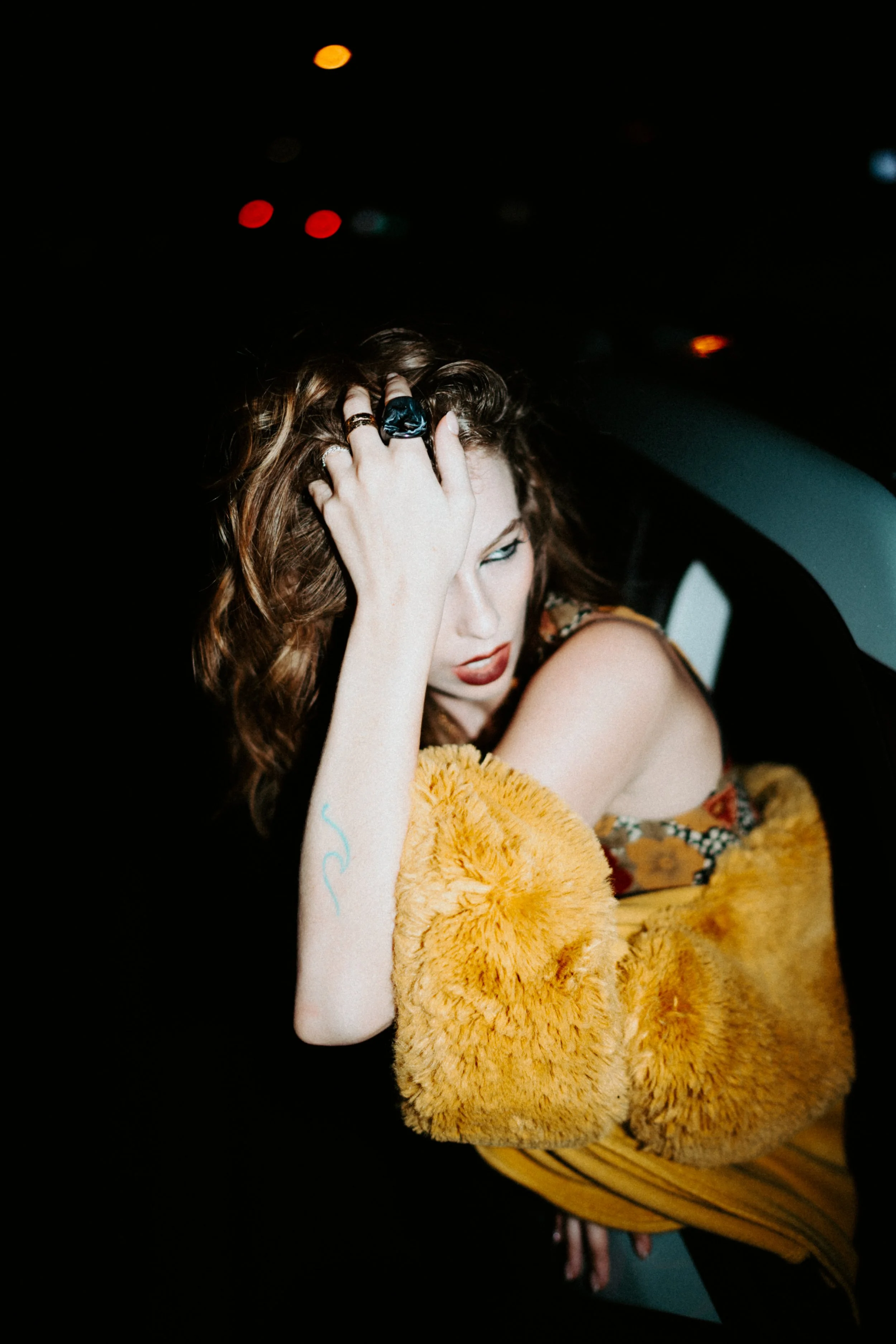 A woman with curly brown hair and fair skin, wearing rings and makeup, is sitting in a car at night. She is wearing a fuzzy orange coat and a floral top, with her hand on her forehead. The background is dark with blurry red and orange lights.