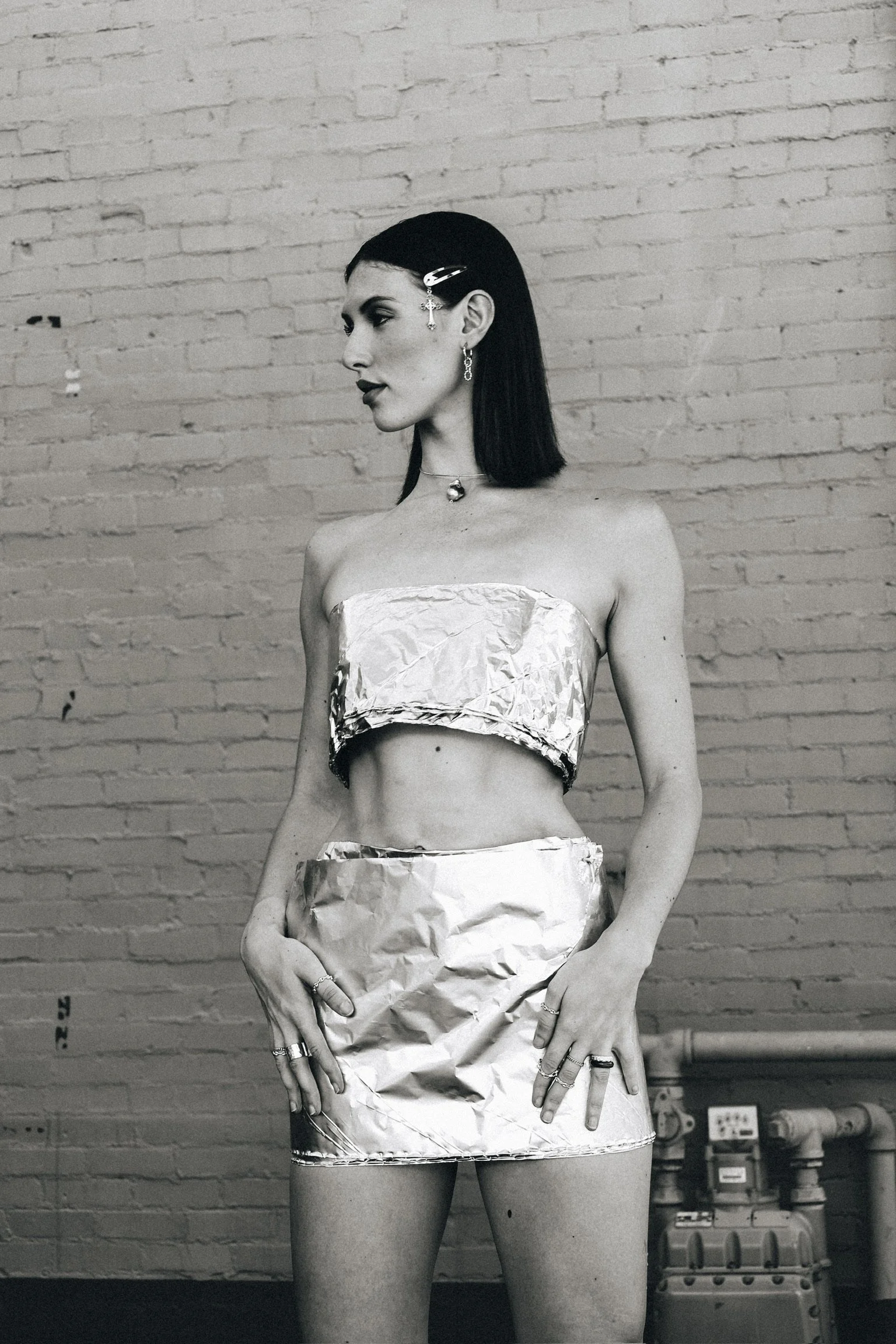 A black and white photo of a woman standing against a brick wall, wearing metallic clothing made of crumpled material, with various jewelry including earrings and necklaces, and a hair clip.