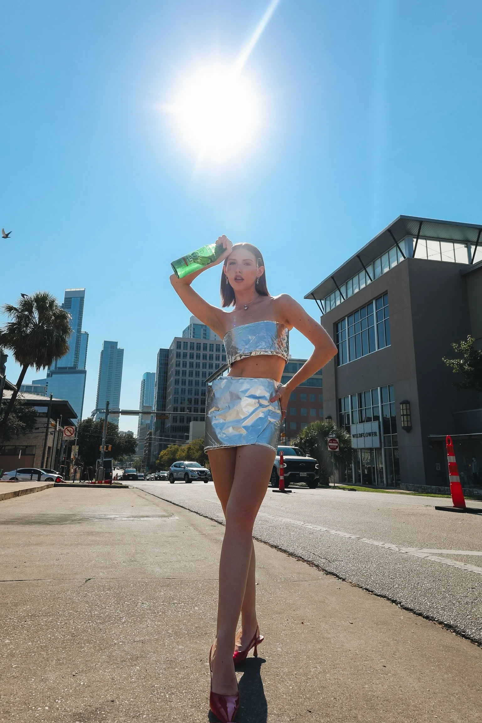 A woman stands barefoot on a city street with skyscrapers behind her. She is holding a bottle to her head, wearing metallic clothing made of reflective material, and red high-heeled shoes. The bright sun is overhead, and a bird is flying in the blue 