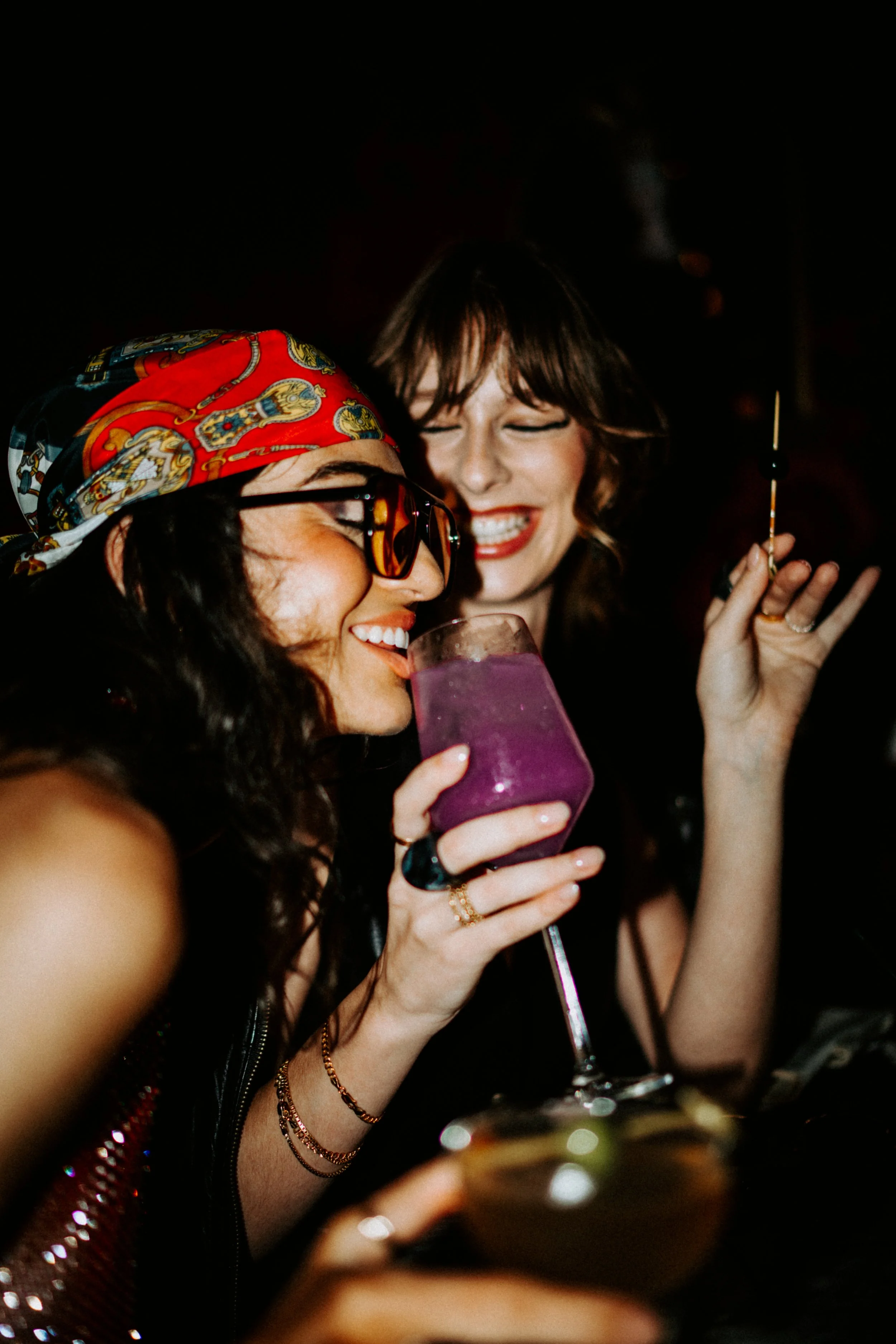 Two women smiling and enjoying drinks at a social event. One woman is wearing a colorful headscarf and sunglasses, holding a purple cocktail, while the other woman has short hair, makeup, and is holding a cigarette.
