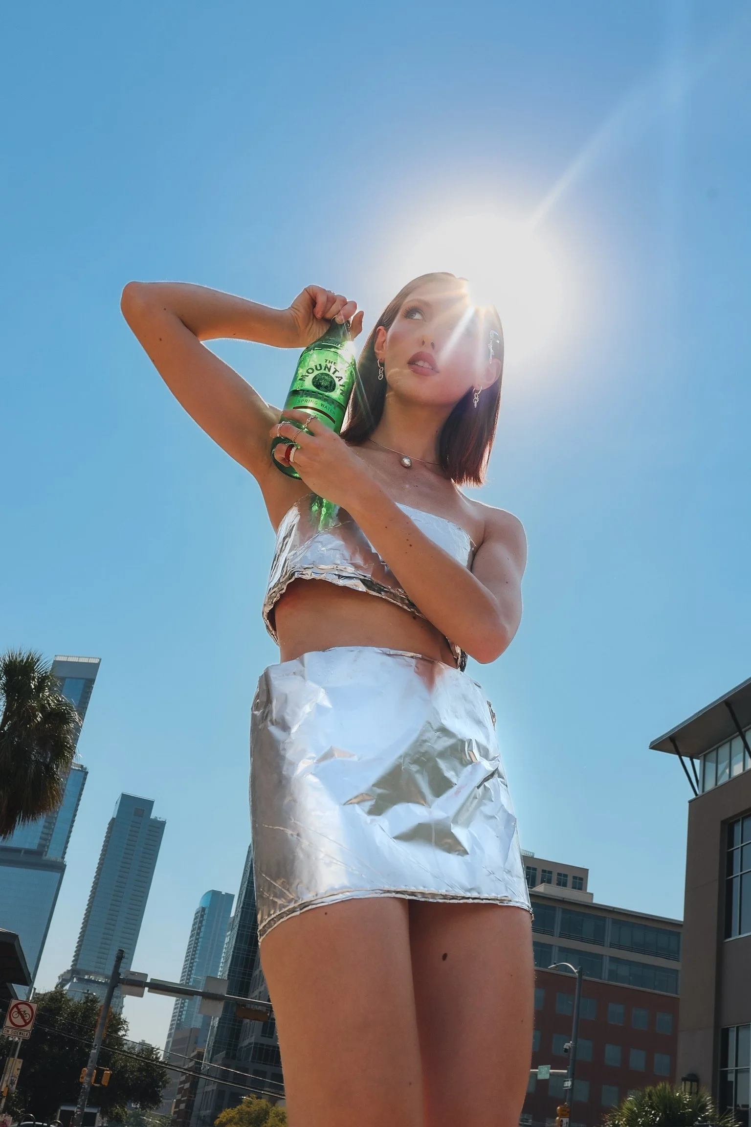 A woman standing outdoors in a cityscape, holding a green water bottle, wearing a silver metallic crop top and matching silver metallic skirt, with the sun shining behind her head against a clear blue sky.