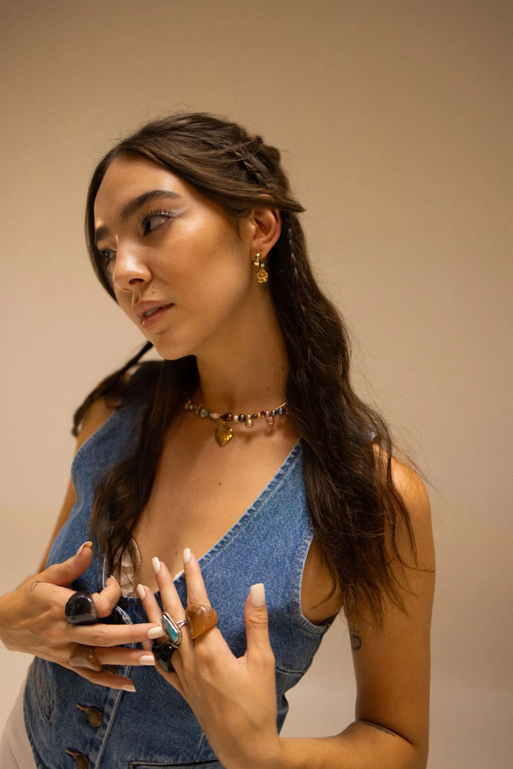 A young woman with long dark hair, wearing gold earrings and a colorful beaded necklace with a gold heart pendant, dressed in a sleeveless blue denim vest, looking to her left, holding the lapel of her vest with her right hand, which is adorned with 
