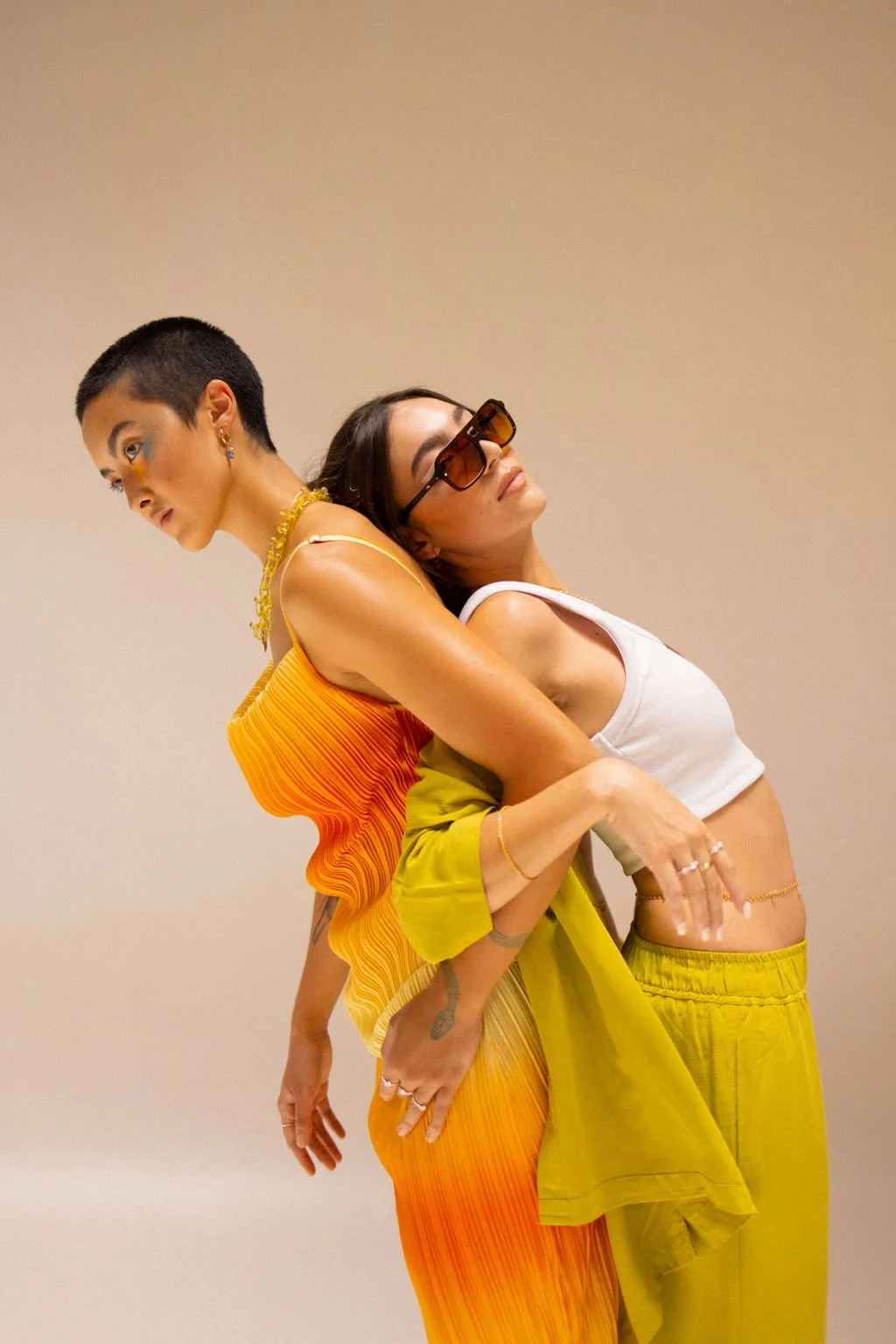 Two women standing closely against a plain wall, wearing colorful fashion-forward clothing. One woman is dressed in a sleeveless orange and yellow gradient dress, with short hair and makeup. The other woman is wearing a white crop top, yellow pants, 