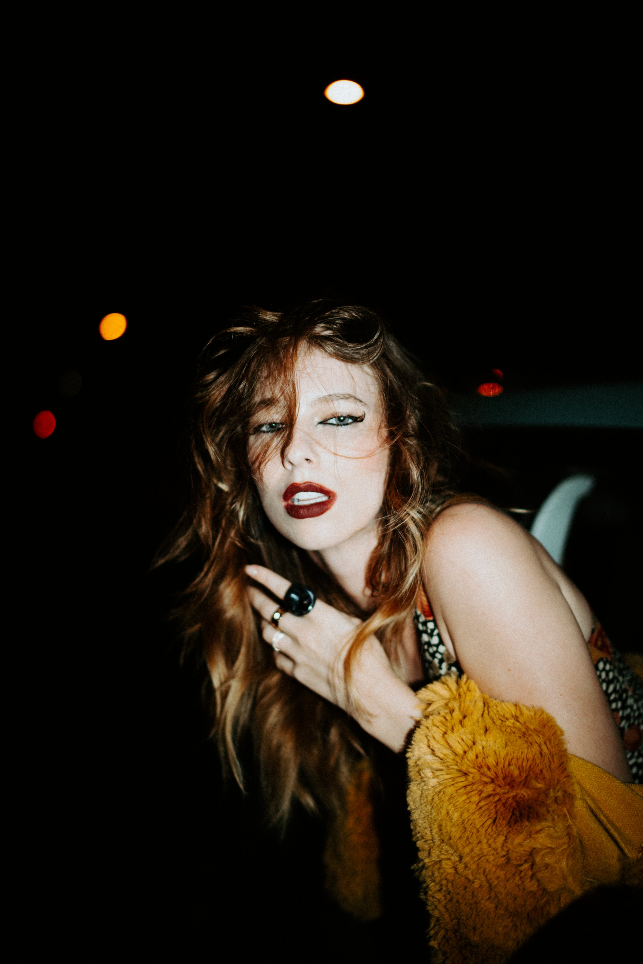 A woman with wavy red hair, wearing dark makeup, including bold lipstick, is posing against a dark background with blurred lights. She is partially wrapped in a yellow fuzzy blanket and has rings on her fingers.