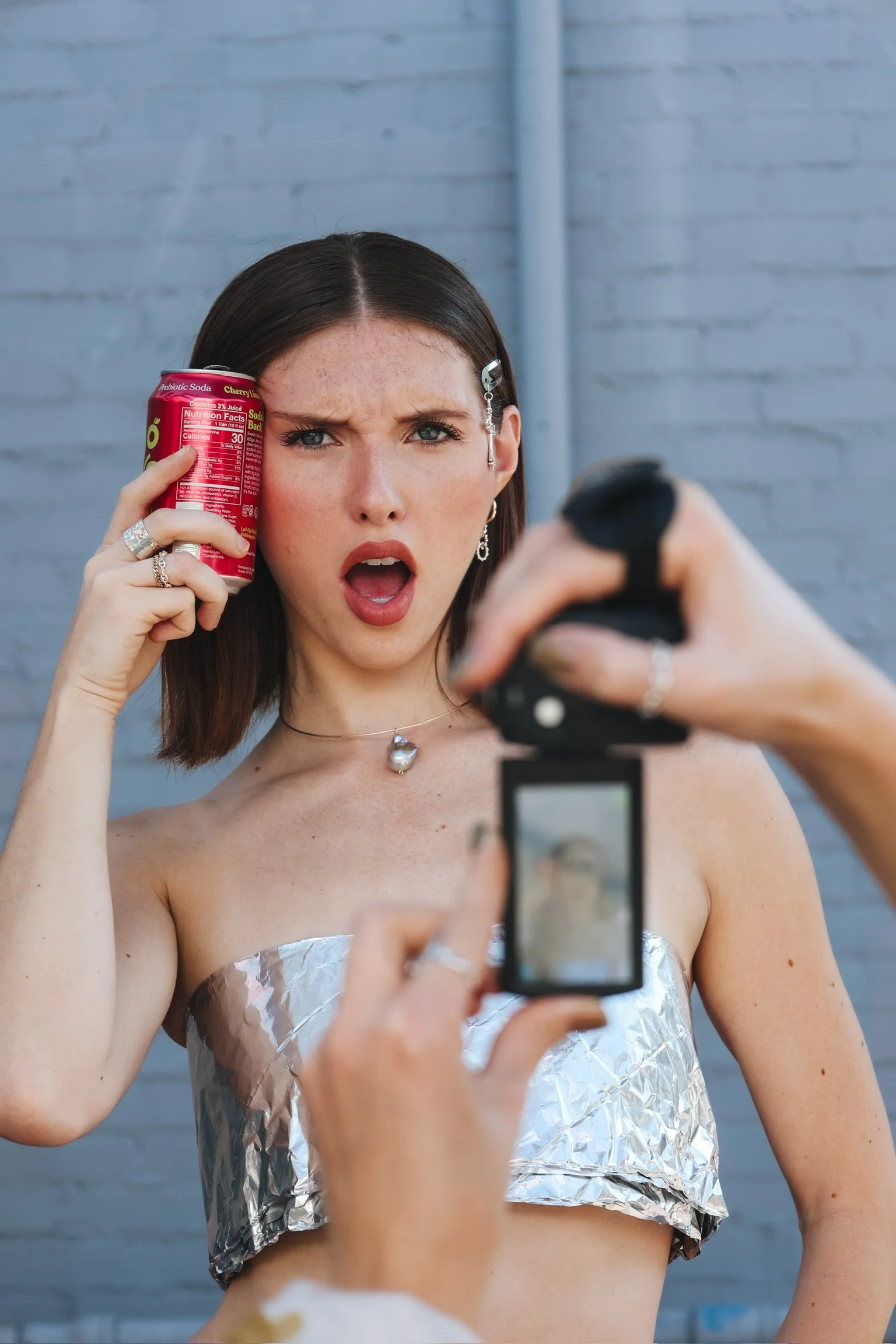 A woman with dark hair and a silver earrings holding a red can of soda up to her face while taking a picture with a smartphone, standing against a gray brick wall.