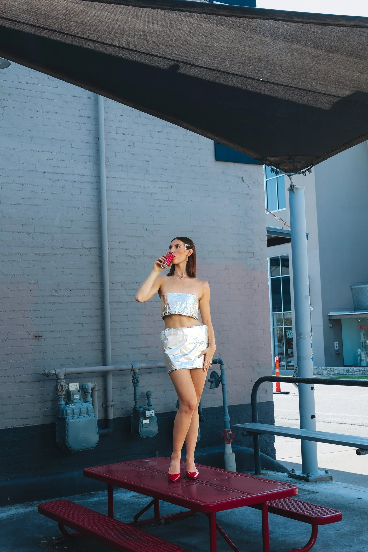 A woman standing on a red picnic table outside, wearing a shiny silver outfit and red shoes, drinking from a can, with a white brick building and blue sky in the background.