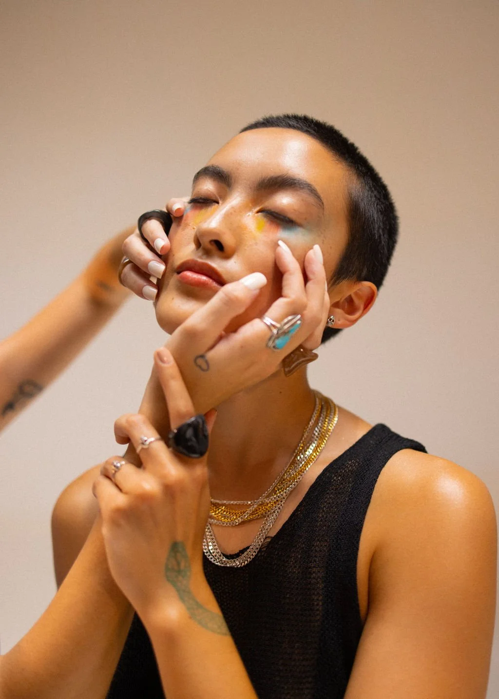 Person with short hair and multiple jewelry pieces having makeup applied around their eyes, with a neutral background.