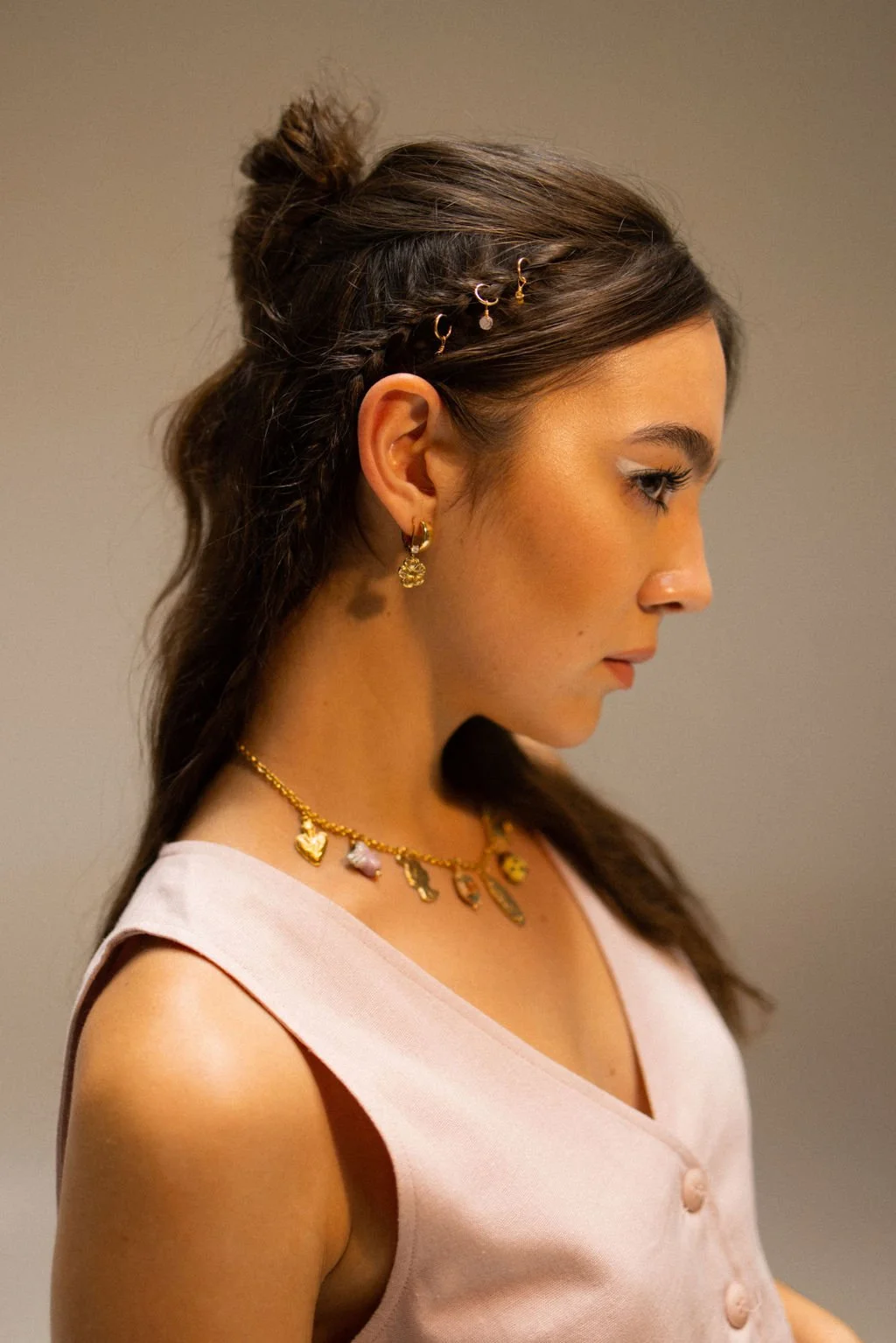 A woman with brown hair styled in a half-up, half-down hairstyle with small gold hairpins. She is wearing gold earrings and a gold necklace with charms. She has a small mole on her neck and is dressed in a pink sleeveless top.