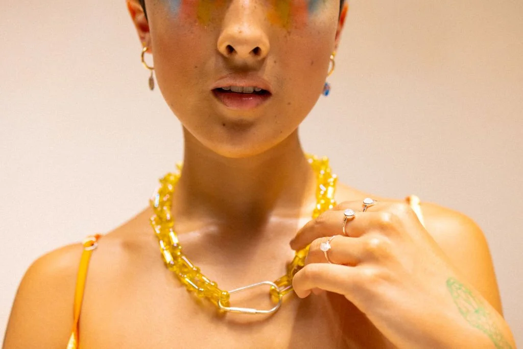 Close-up of a woman wearing yellow jewelry, including a necklace and rings, with earrings, touching her necklace with her hand, against a plain background.