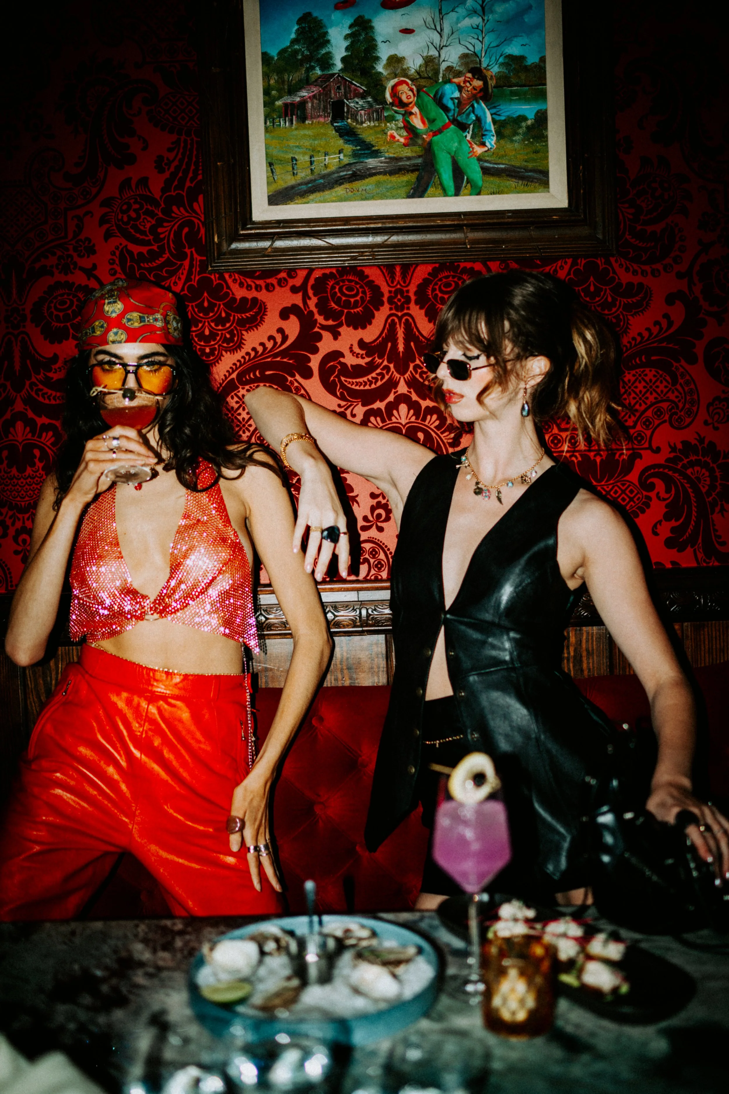 Two women dressed in colorful, fashionable outfits at a party with a red patterned wall and a painting of a rural scene behind them. One woman is wearing a red satin top, red pants, sunglasses, and a patterned headscarf, while the other is in a black