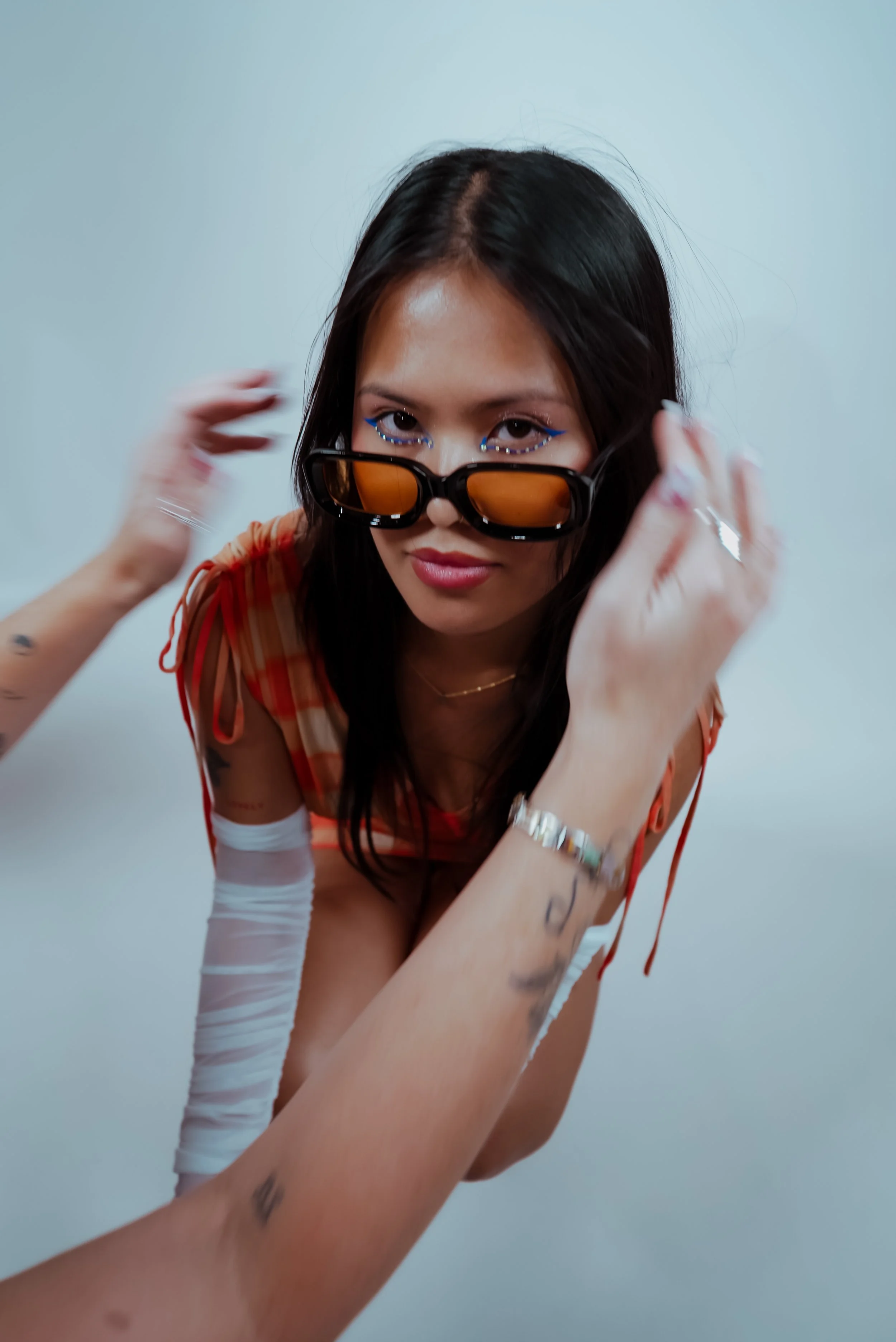 A woman with long dark hair, wearing orange-tinted sunglasses, blue eye makeup, and a checkered orange and red top. She is adjusting her glasses and looking at the camera with a slight smile, against a plain light-colored background.