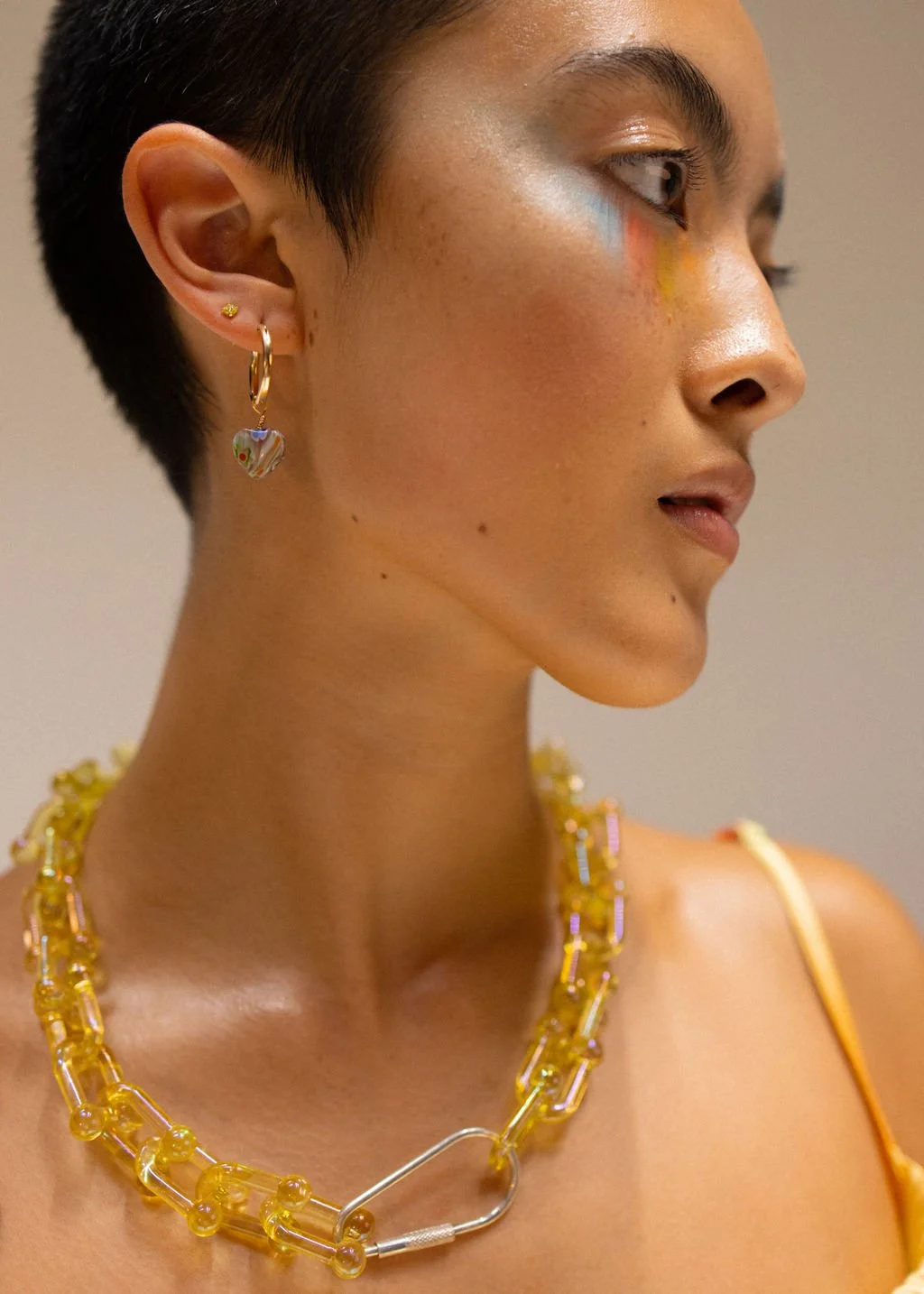 Close-up side profile of a woman with short dark hair, wearing colorful earrings and a bold yellow chain necklace.
