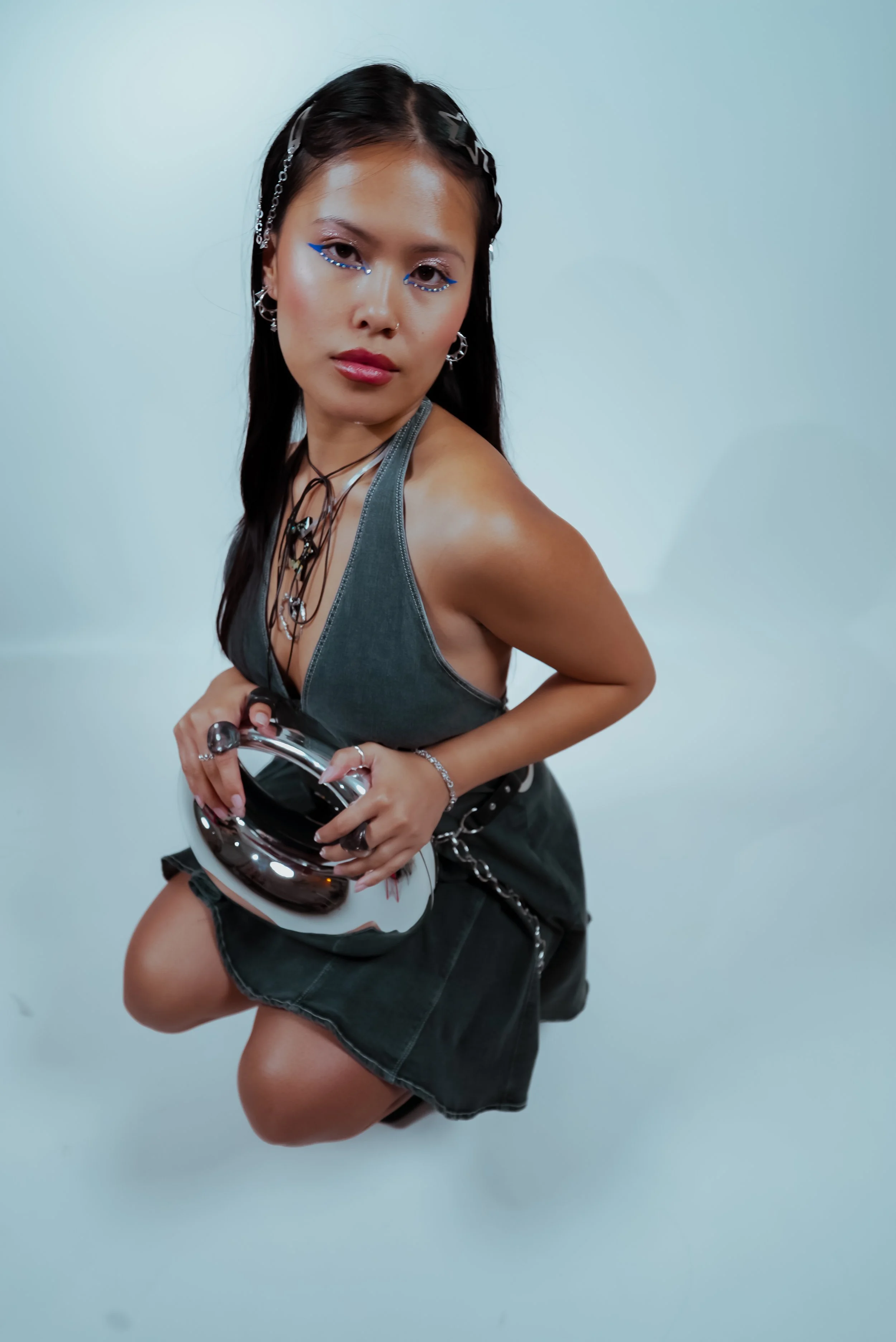 A young woman with dark hair styled with hair clips, wearing makeup with blue eyeliner, silver jewelry, a tank top, and a leather skirt, holding a helmet against a plain background.