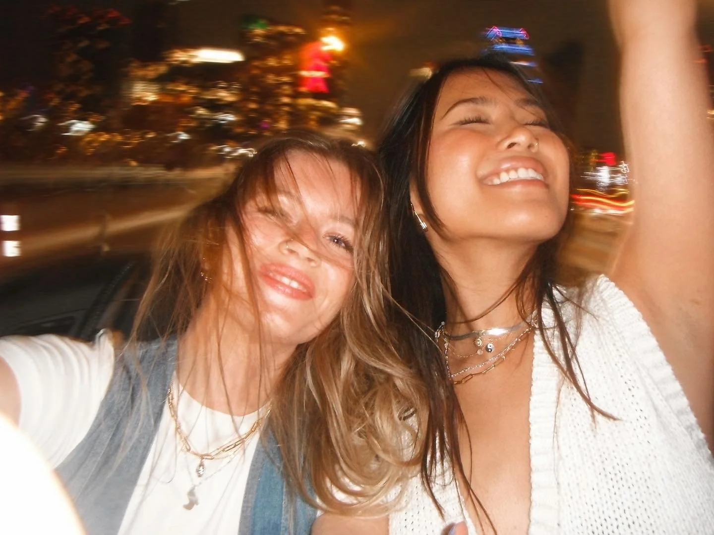 Two women taking a selfie at night with blurred city lights in the background, smiling and enjoying themselves.