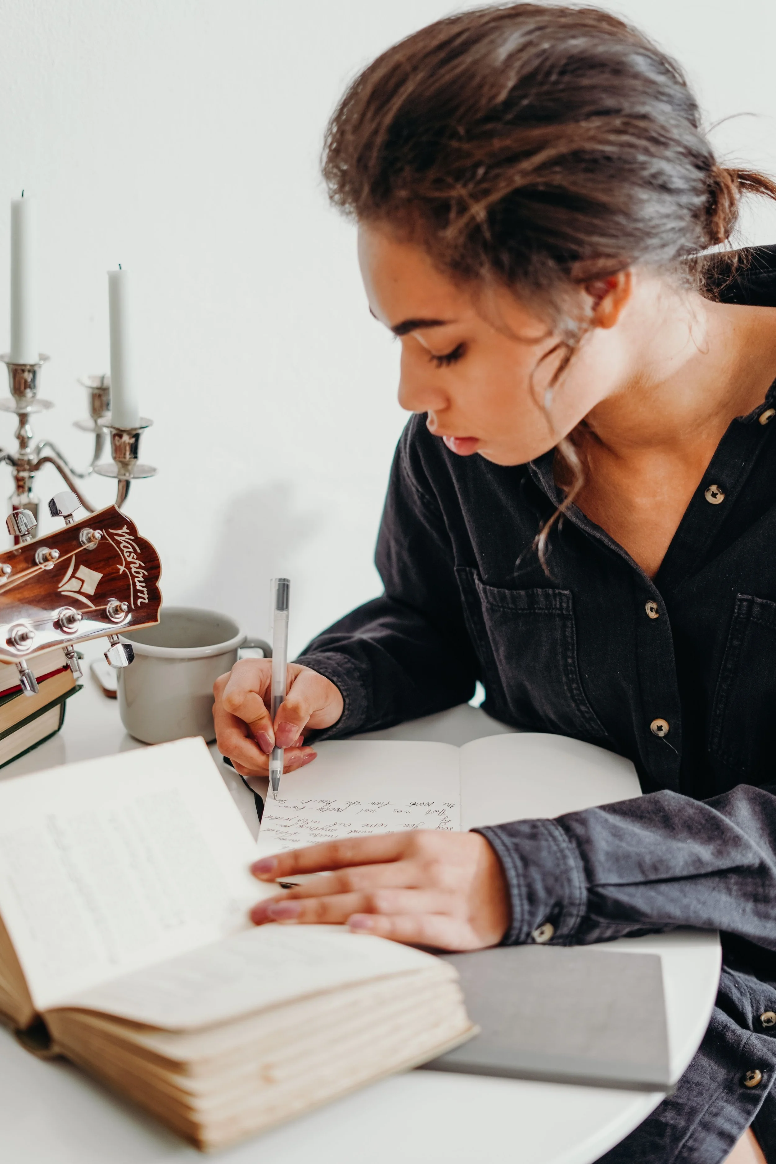 A woman with brown hair styled in loose waves, wearing a dark jacket, is sitting at a white table, writing in a notebook with a pen. A guitar headstock is visible on the left side of the image, along with a candlestick holder and a mug on the table.