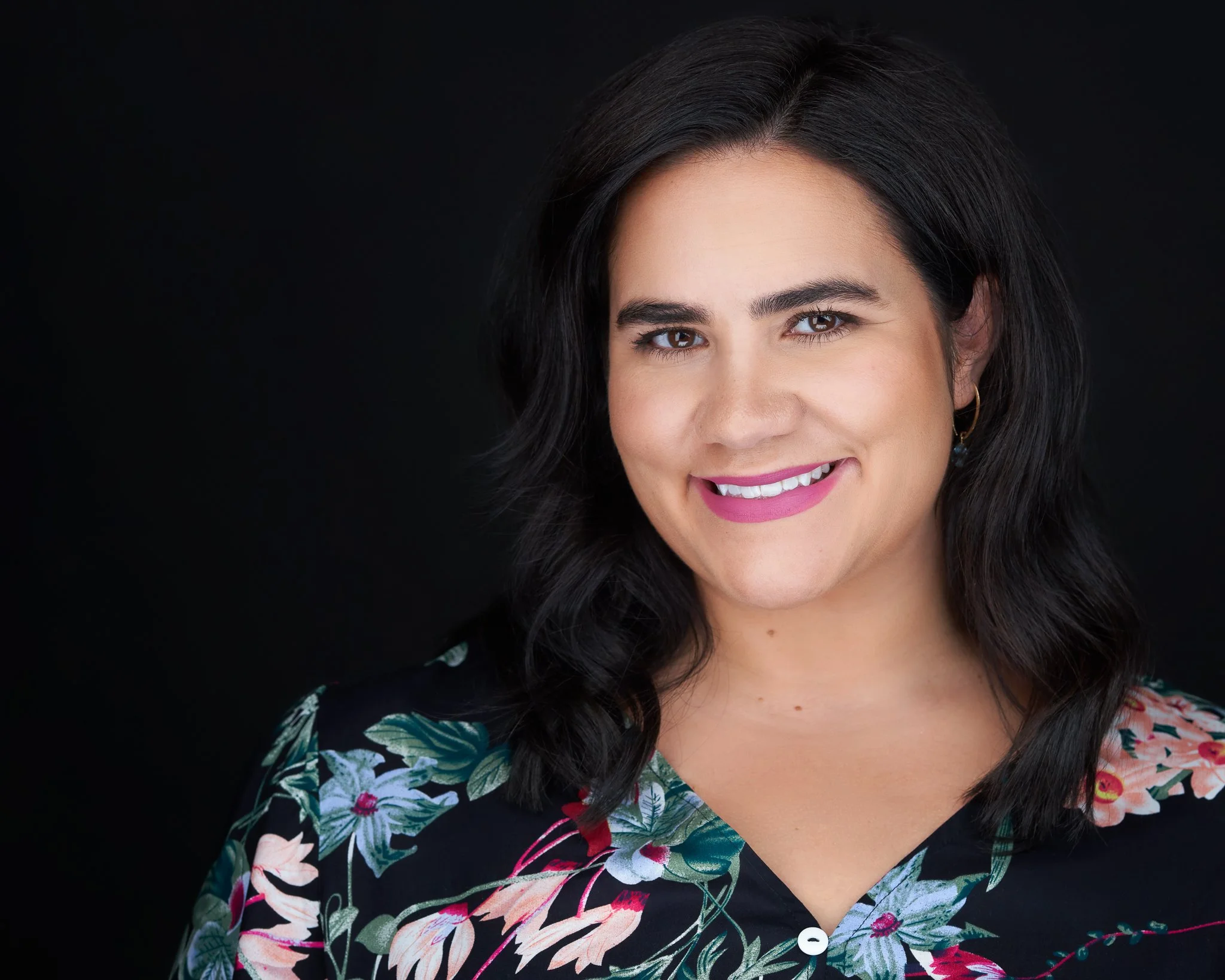 Close-up of a woman with dark hair, smiling, wearing a floral top with a black background.