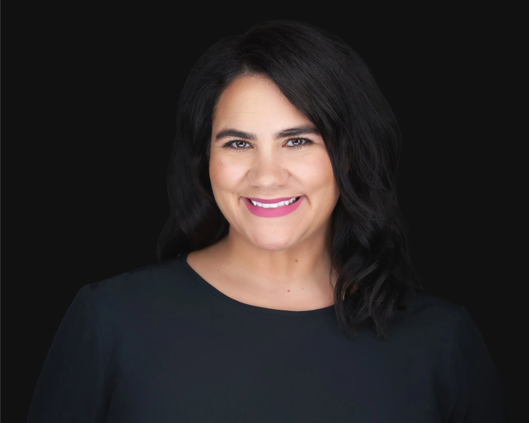 Close-up portrait of a woman with dark hair, wearing a black top, smiling against a black background.