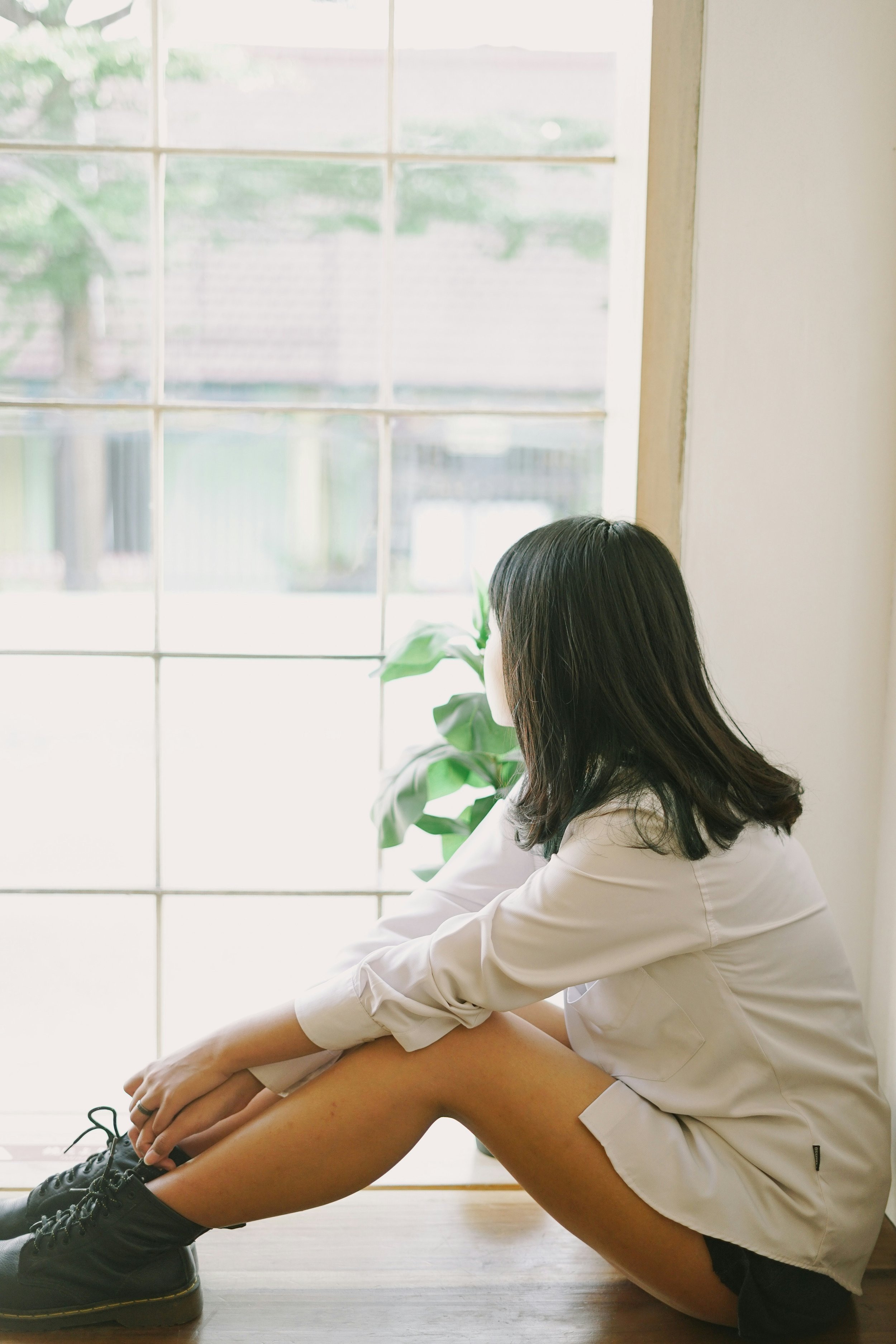 A woman sitting on the floor in front of a large window, tying her shoe, with another person sitting in front of her, their bare legs visible. The scene suggests a relaxed, casual moment indoors.