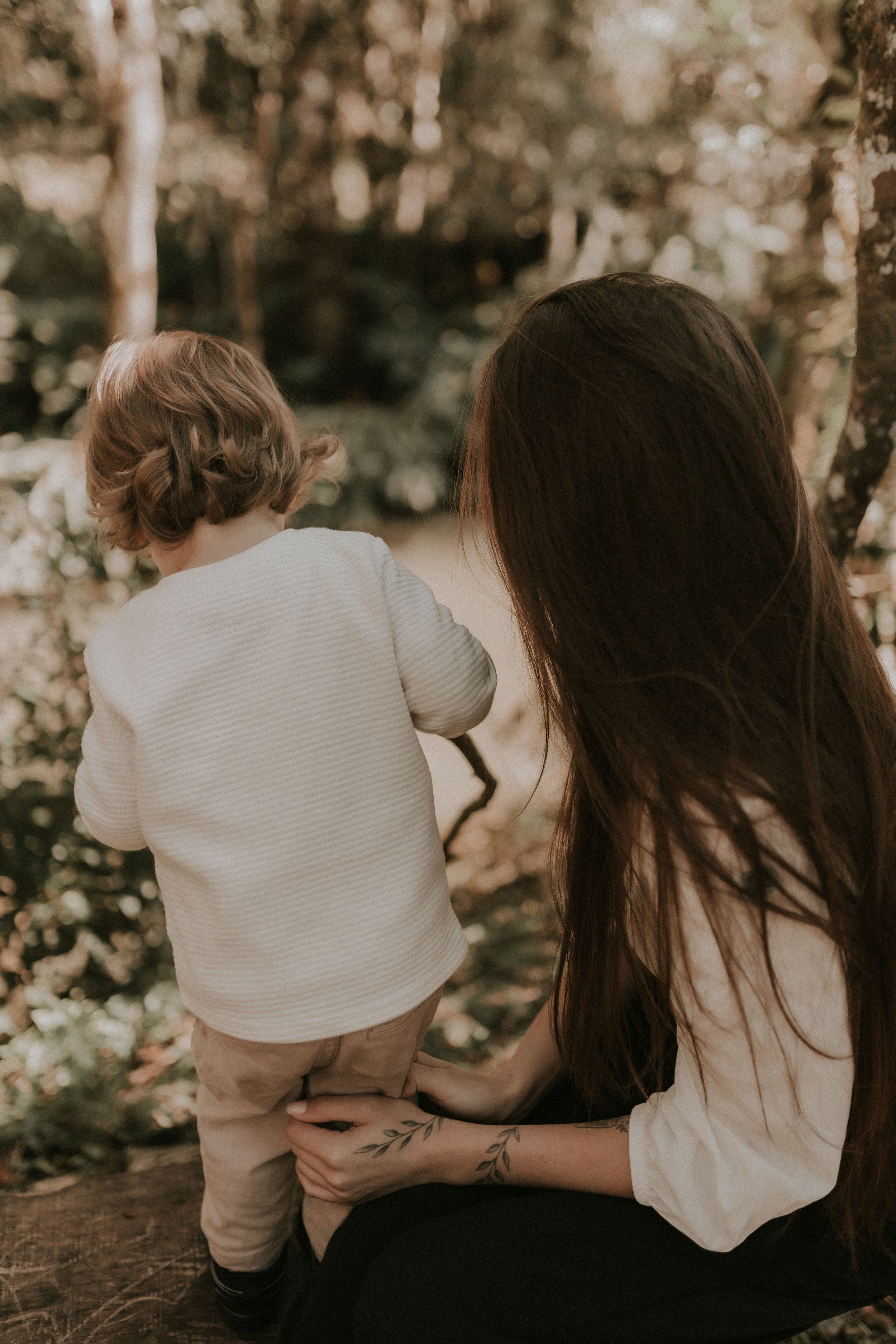 A woman with long dark hair and tattoos on her arm is holding a toddler with curly hair, in a wooded outdoor setting.