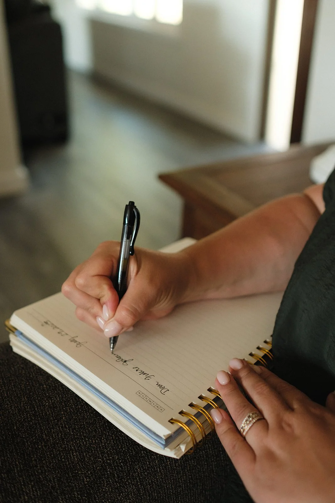 A person writing in a spiral notebook with a black pen, sitting on a dark surface, with a wedding ring on their finger and a piece of furniture nearby.