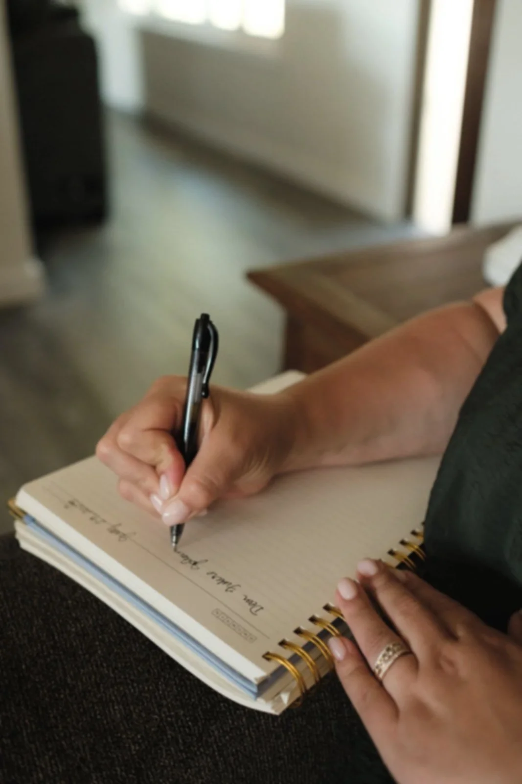 A person writing in a spiral-bound notebook with a black pen, sitting on a brown couch in a room with a window in the background.