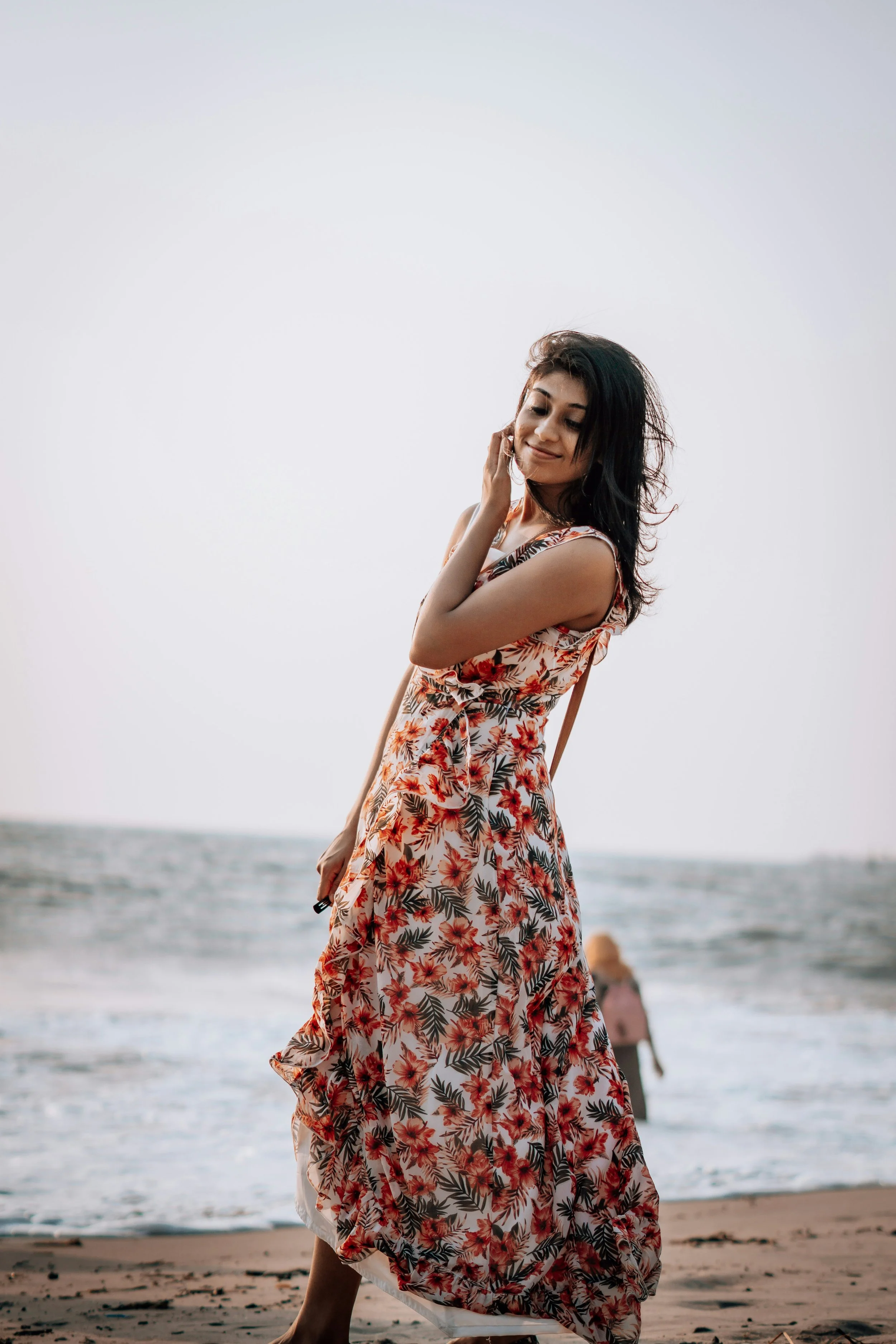 A woman in a sleeveless floral dress standing on the beach near the ocean, with her eyes closed and a gentle smile, in front of a bright sky and a person in the background.