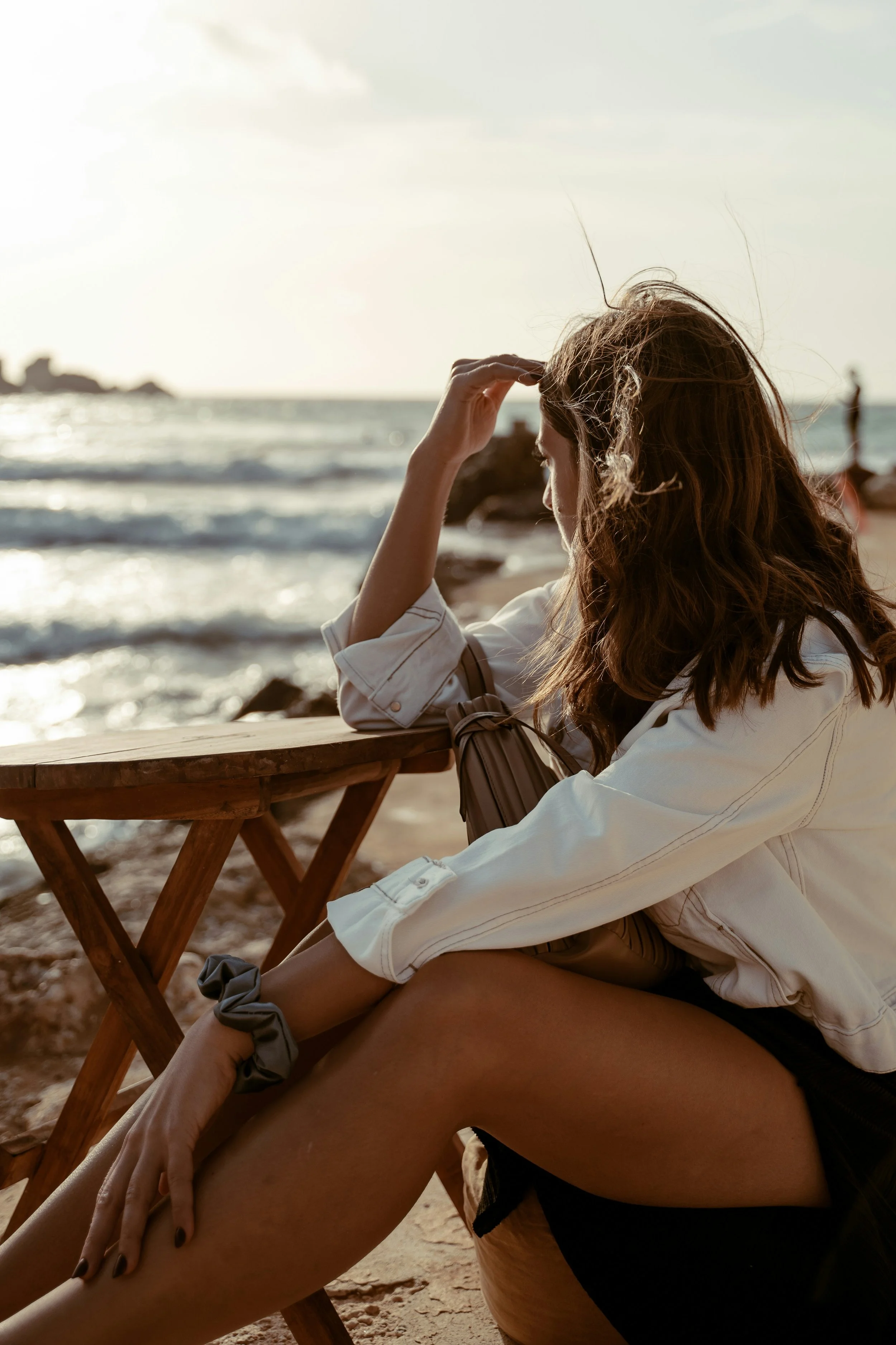 A woman with long wavy hair sitting at a beachside with a woman in black shorts and a gray scrunchie on her wrist. The woman is resting her chin on her hand while looking at the ocean, with rocky formations and other people in the background.