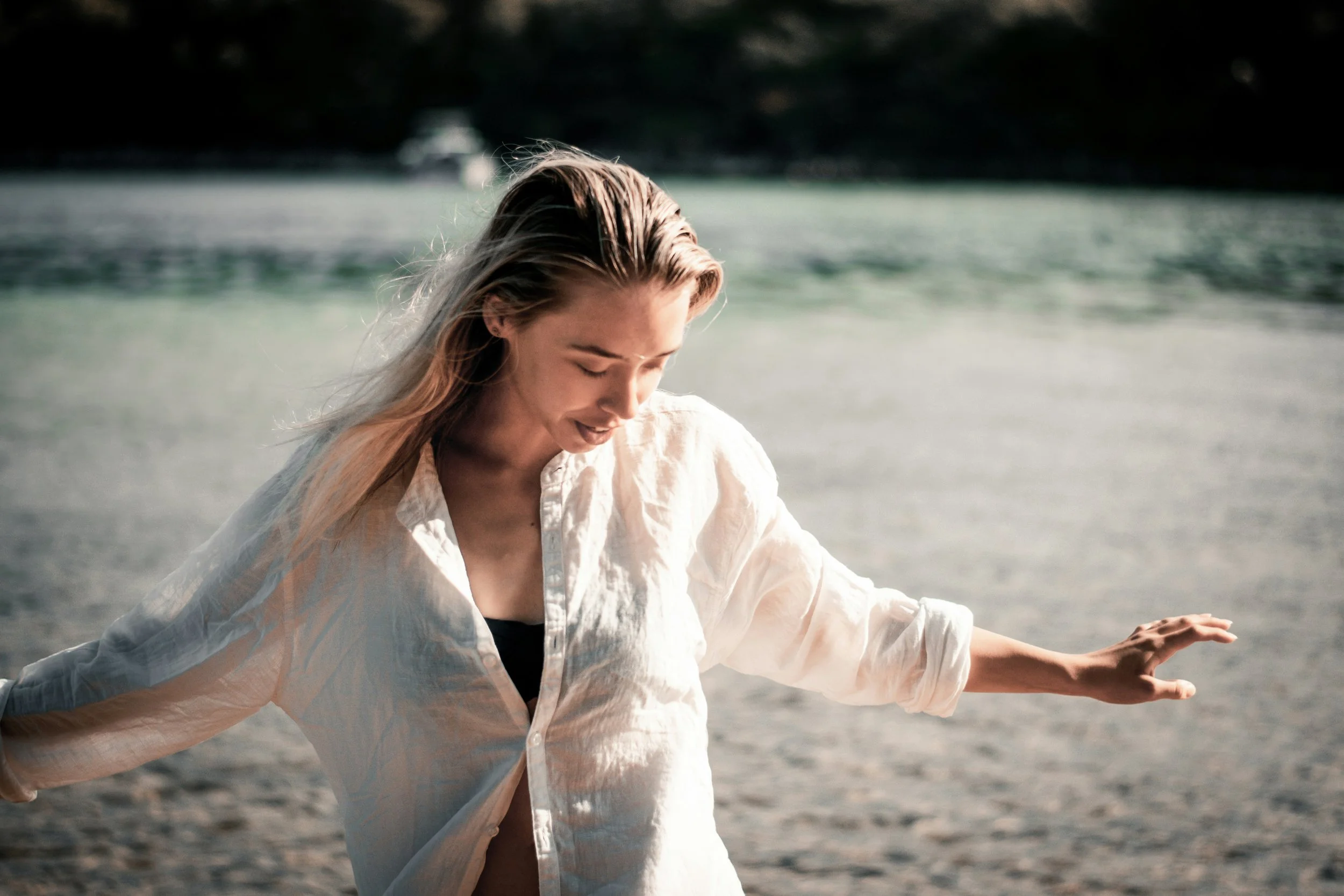 A woman with blonde hair wearing a white shirt, standing on a beach with water in the background, looking down with a peaceful expression.