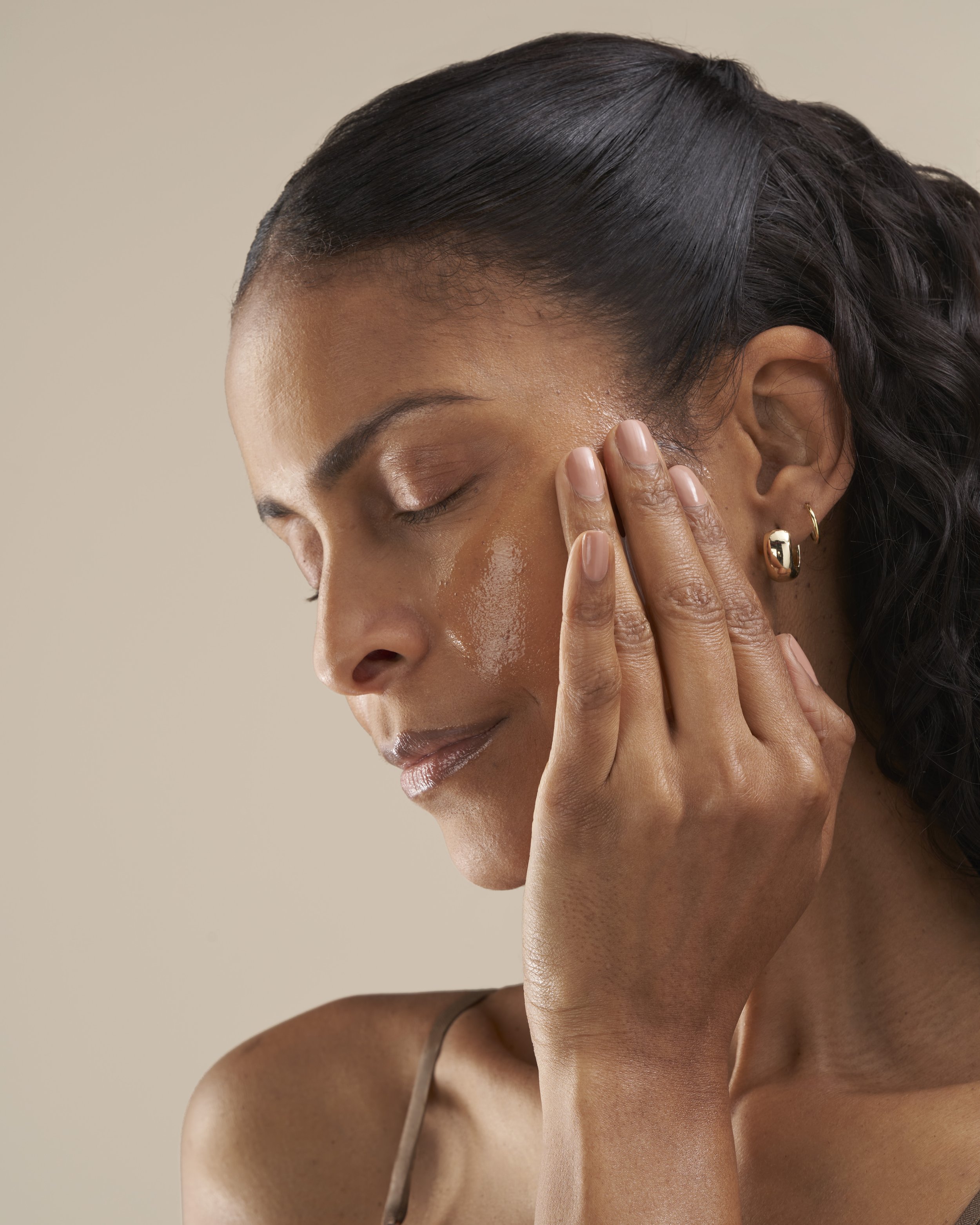 A woman with closed eyes has her hand gently touching her temple, with smooth, shiny black hair styled in waves, wearing gold hoop earrings and a thin strap top, against a beige background.
