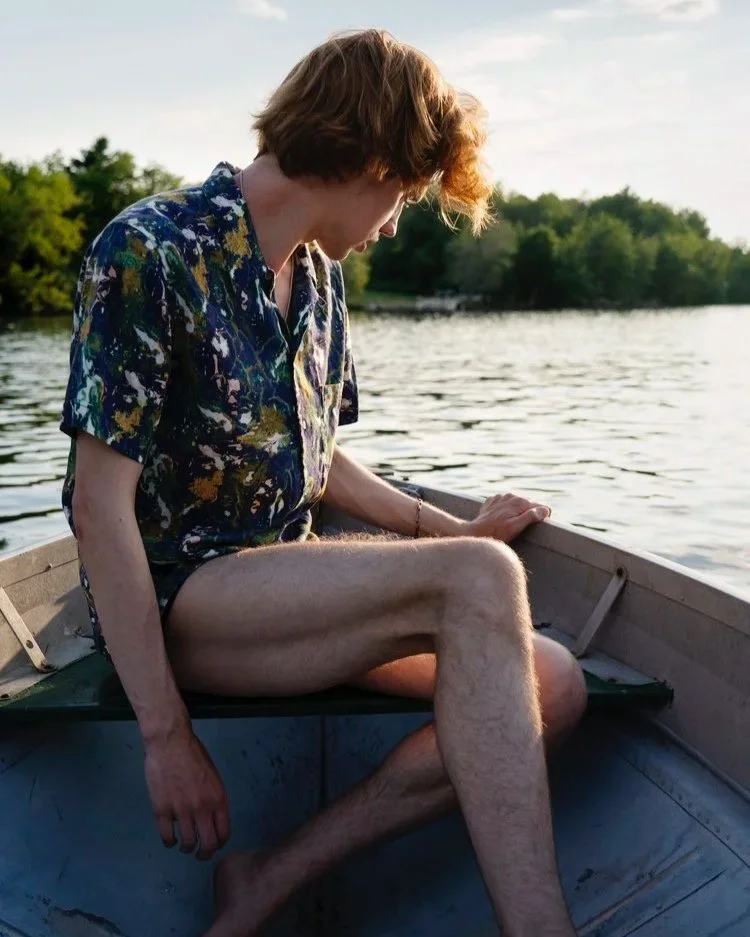 A person with shoulder-length red hair sitting in a small boat on a lake, wearing a colorful patterned shirt and shorts, with trees and water visible in the background during sunset.
