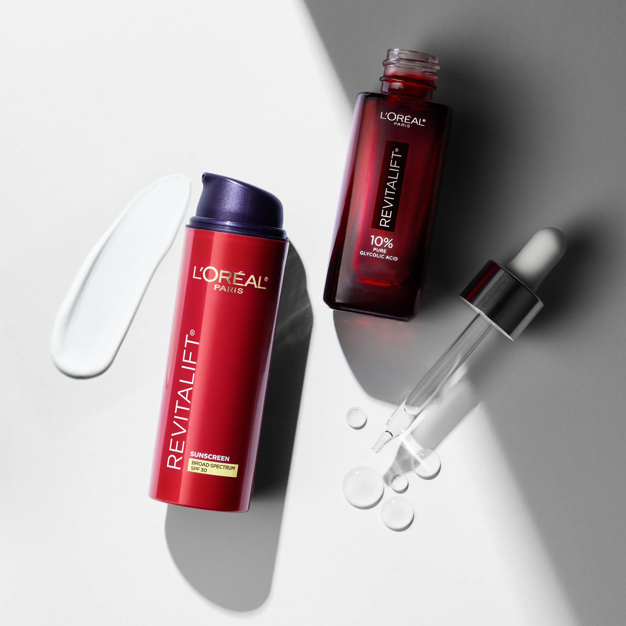 A red bottle of L'Oréal Paris Revitalift 10% Pure Glycolic Acid, with a white and purple applicator and drops of liquid on a white and gray background.