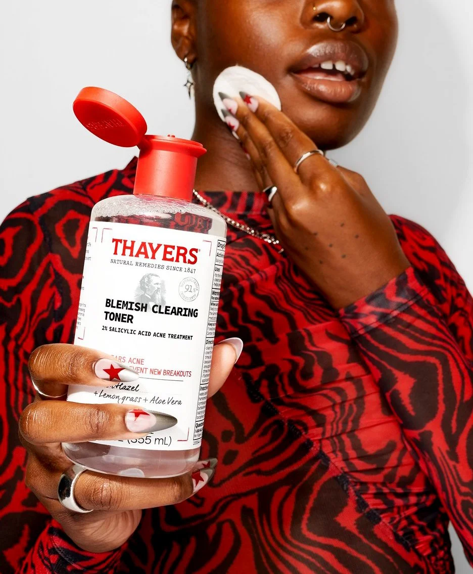 Person with dark skin applying Thayers Blemish Clearing Toner to their face using a cotton pad, holding a bottle of the toner with a red cap and white label. They wear a red and black patterned shirt, have a nose ring, several rings, and long nails w