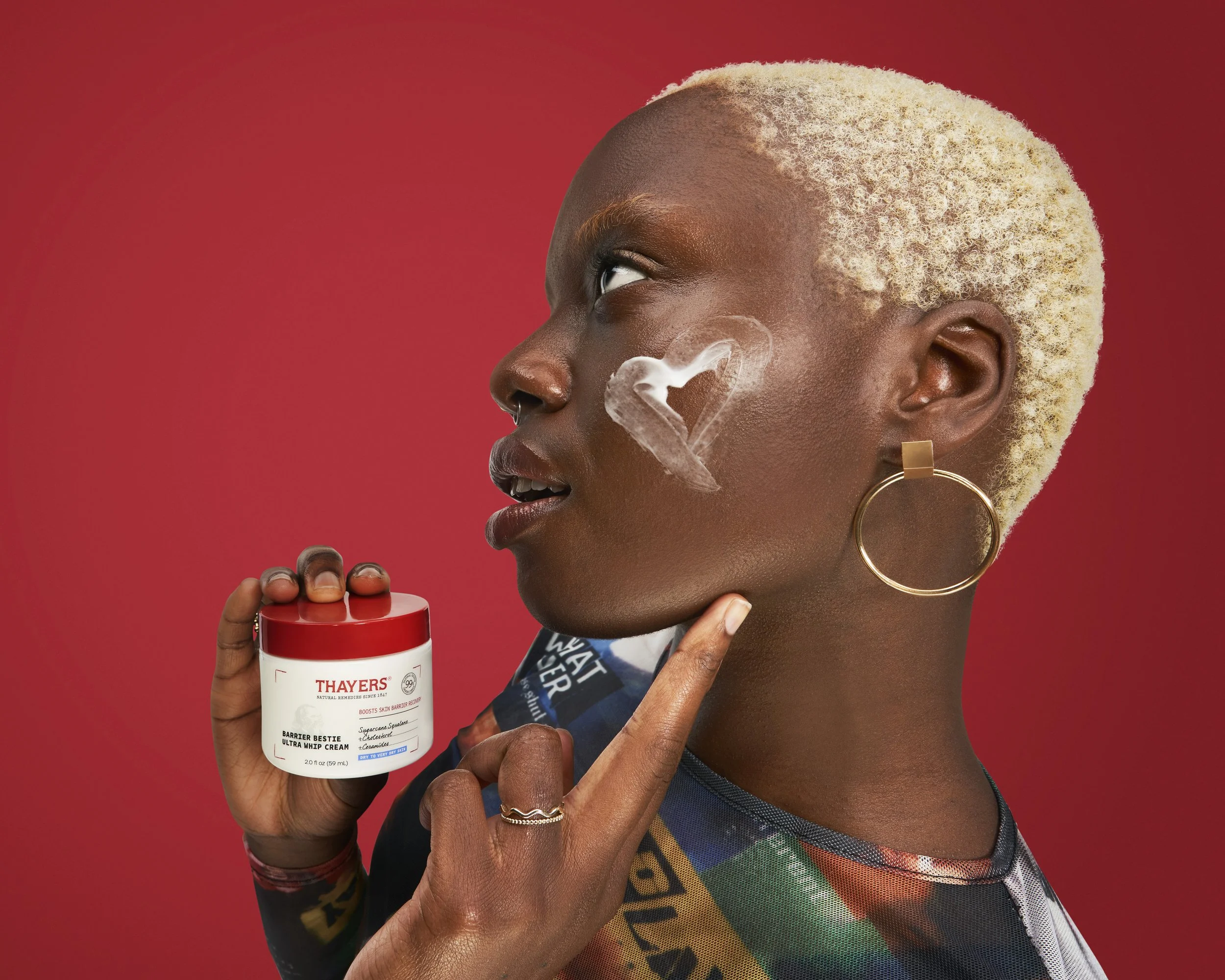 A woman with blonde curly hair, wearing large hoop earrings, holding a container of Thayers Ultra White Cream, with steam forming a heart shape near her face, against a solid red background.