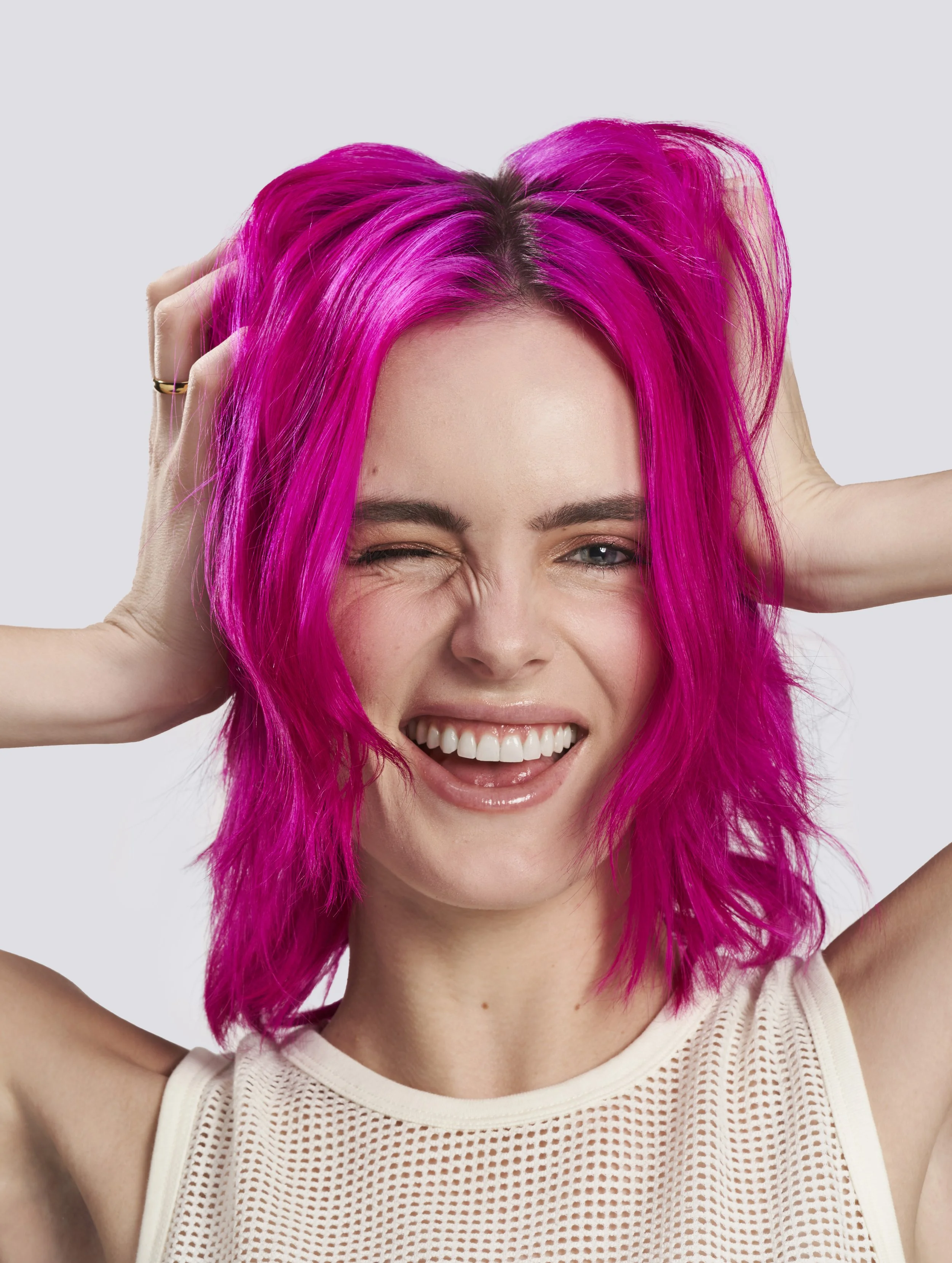 A woman with bright pink hair, winking and smiling, wearing a white sleeveless mesh top and holding her hair with her hands.