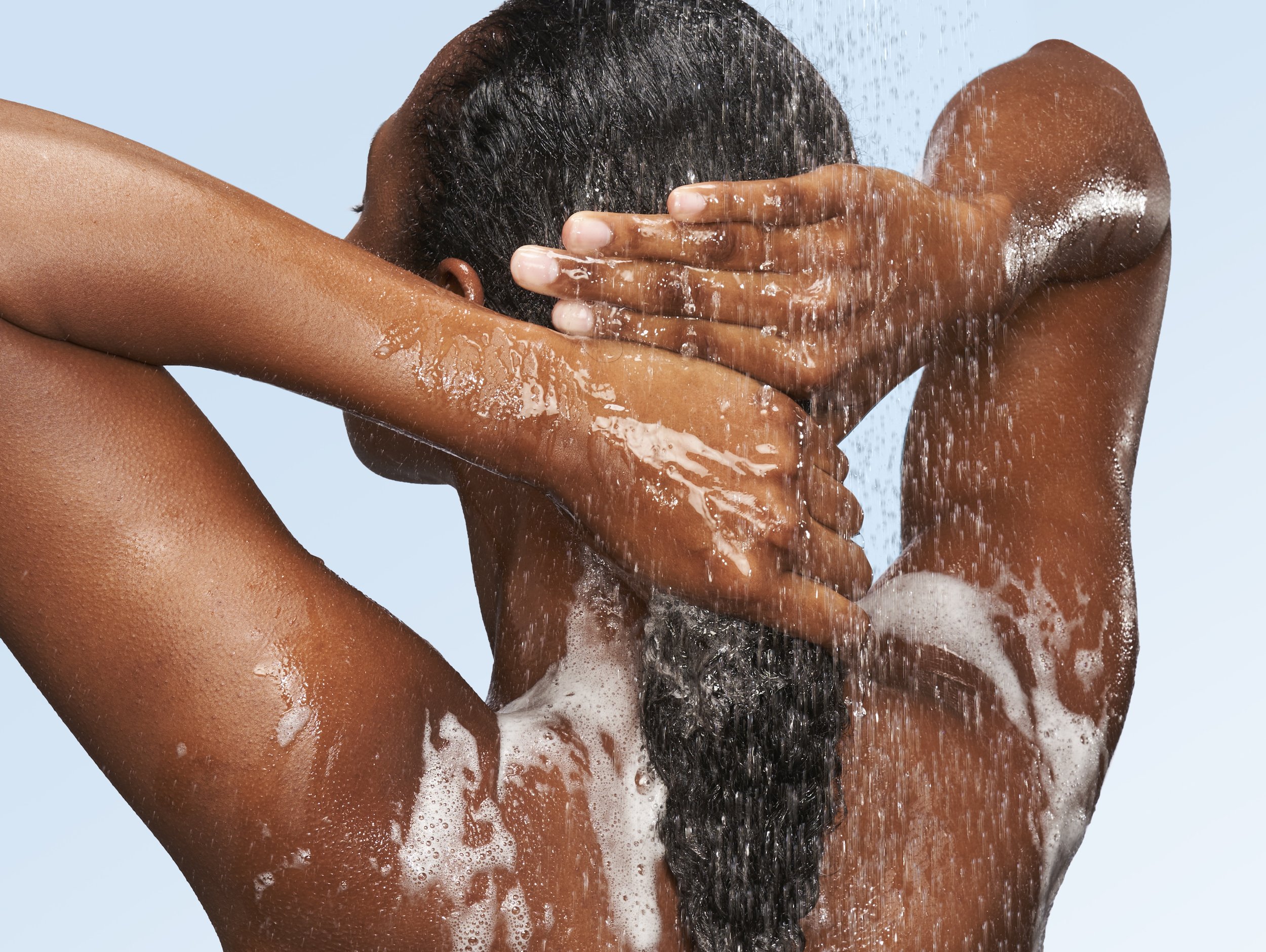 A person with dark hair washing their head under a shower with water and soap suds.