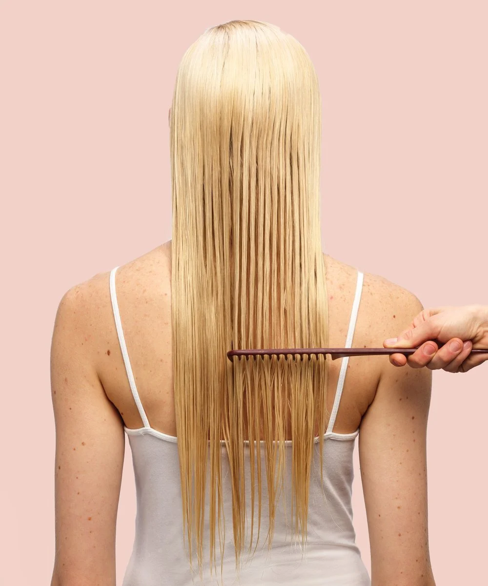 A person with long, straight blonde hair being combed with a brown comb against a pink background.