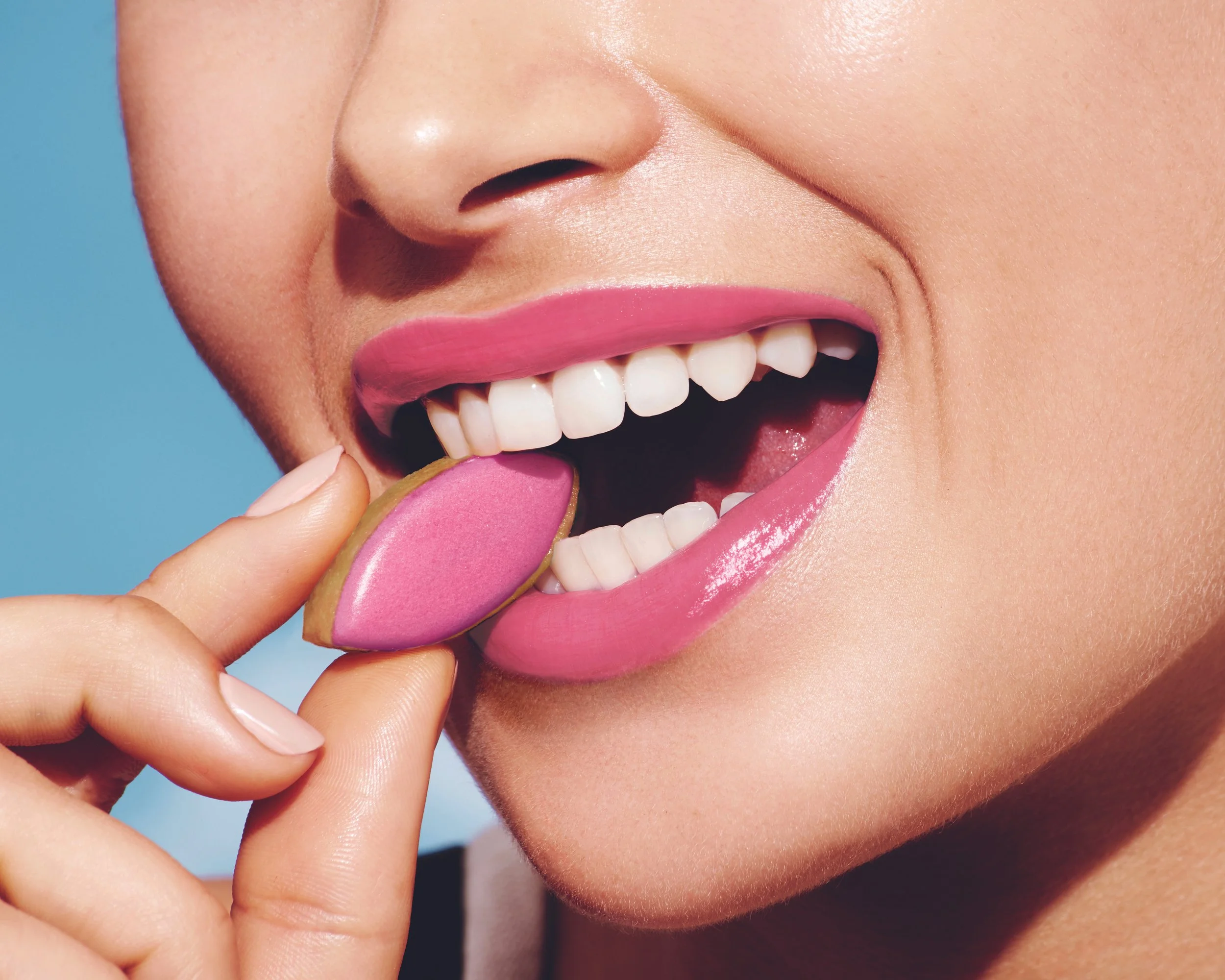 A close-up of a person smiling with pink lipstick, exposing white teeth, holding a pink macaroon near their mouth.