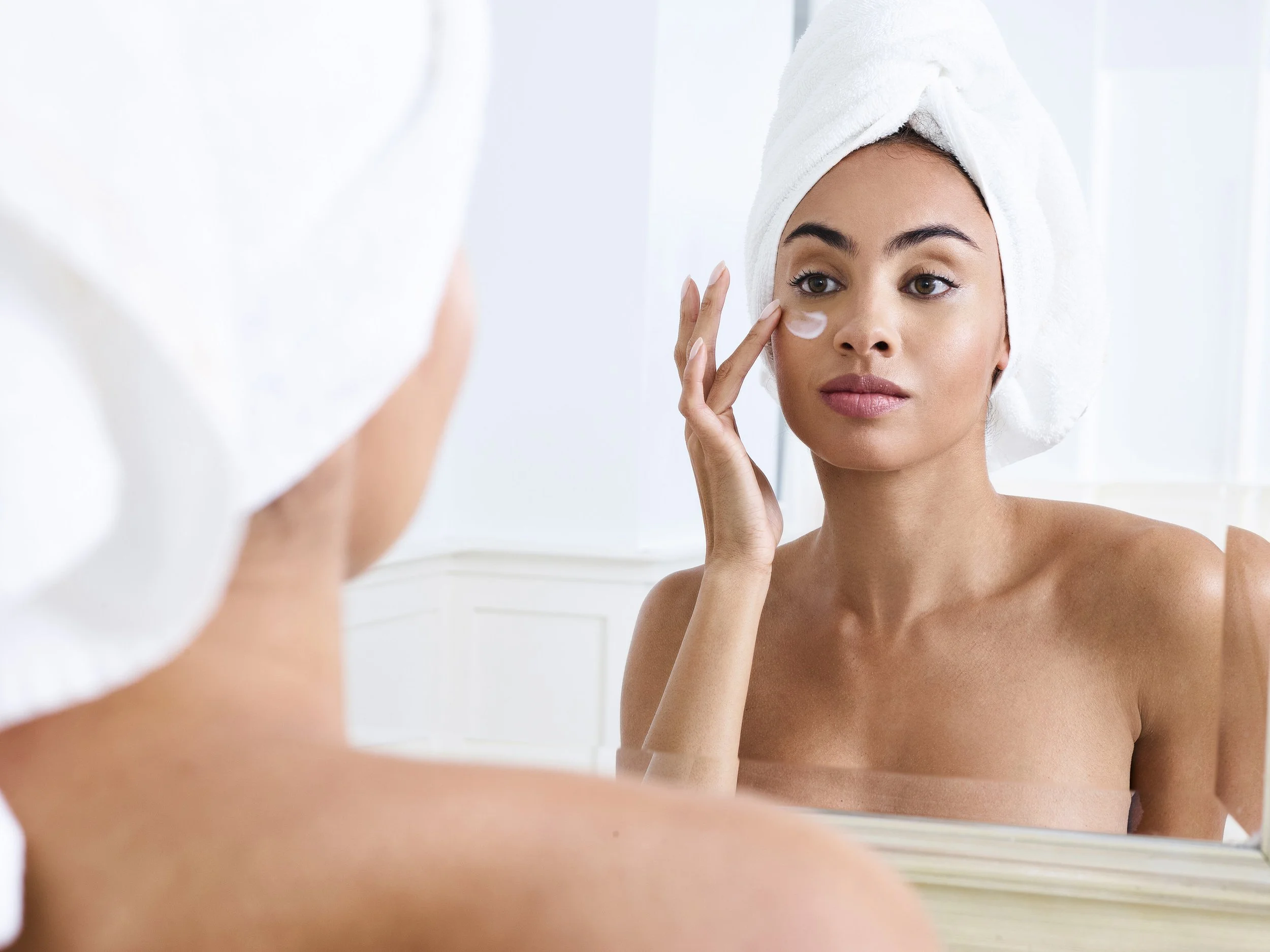 A woman with a towel wrapped around her head looking at herself in a mirror before applying eye cream.