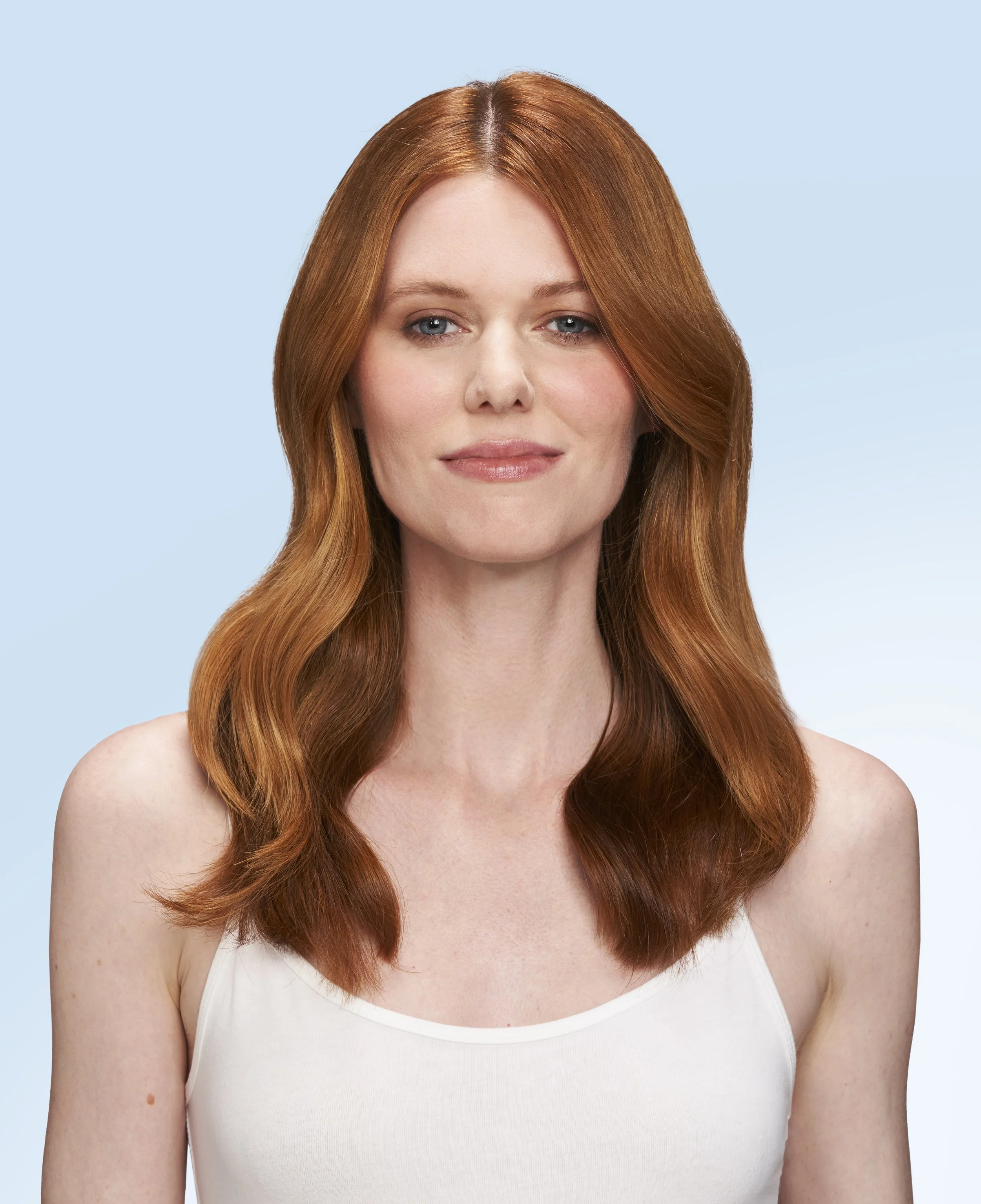 Close-up of a woman with long, red hair, fair skin, and wearing a white tank top, smiling softly against a light blue background.