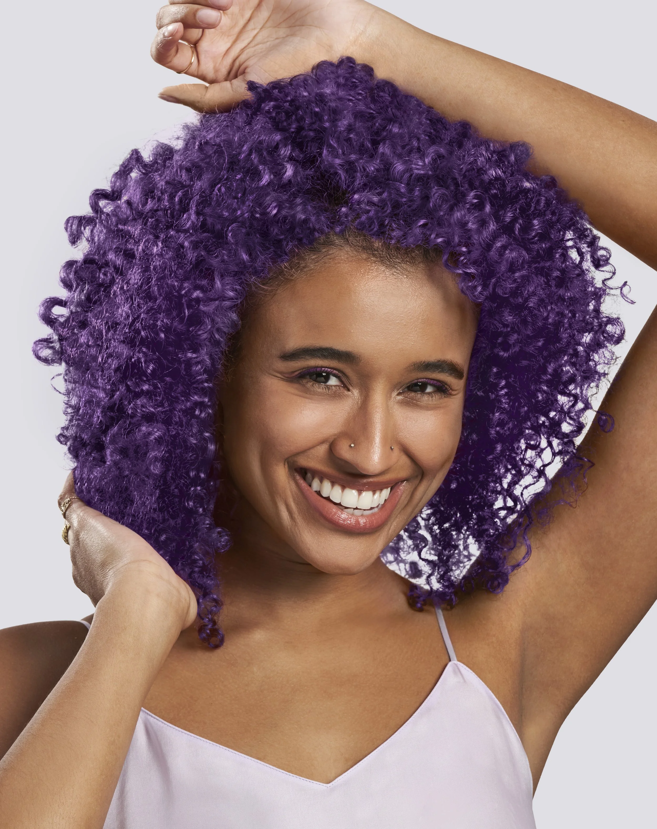 A woman with purple curly hair, smiling, wearing a light purple top, resting her hand on her head.
