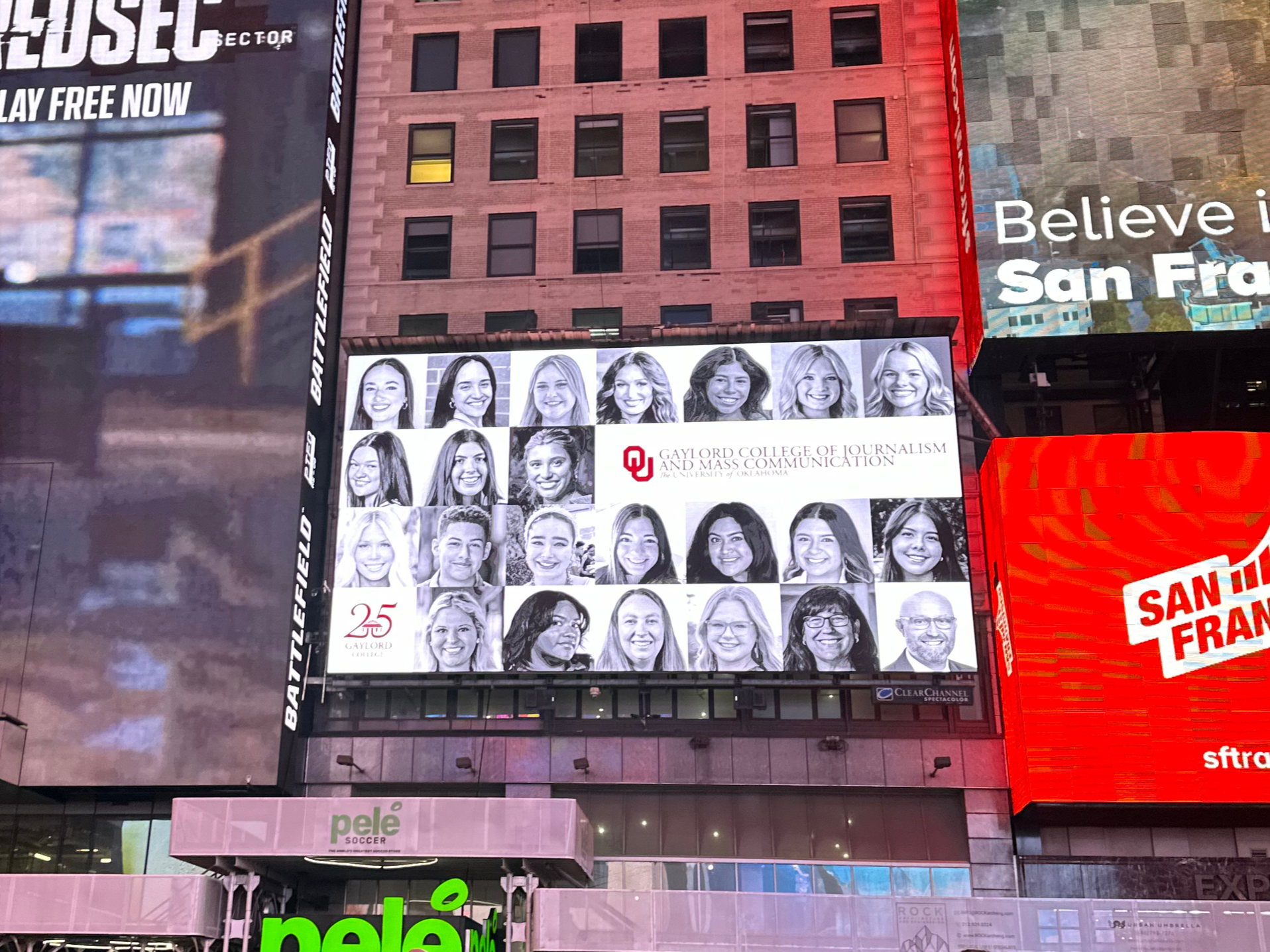 Large digital billboard displaying black and white portraits of diverse individuals on the facade of a building, with the logo of Gaylord College of Journalism and Mass Communication at the University of Oklahoma, and other colorful signs and advertisements in the surrounding area.