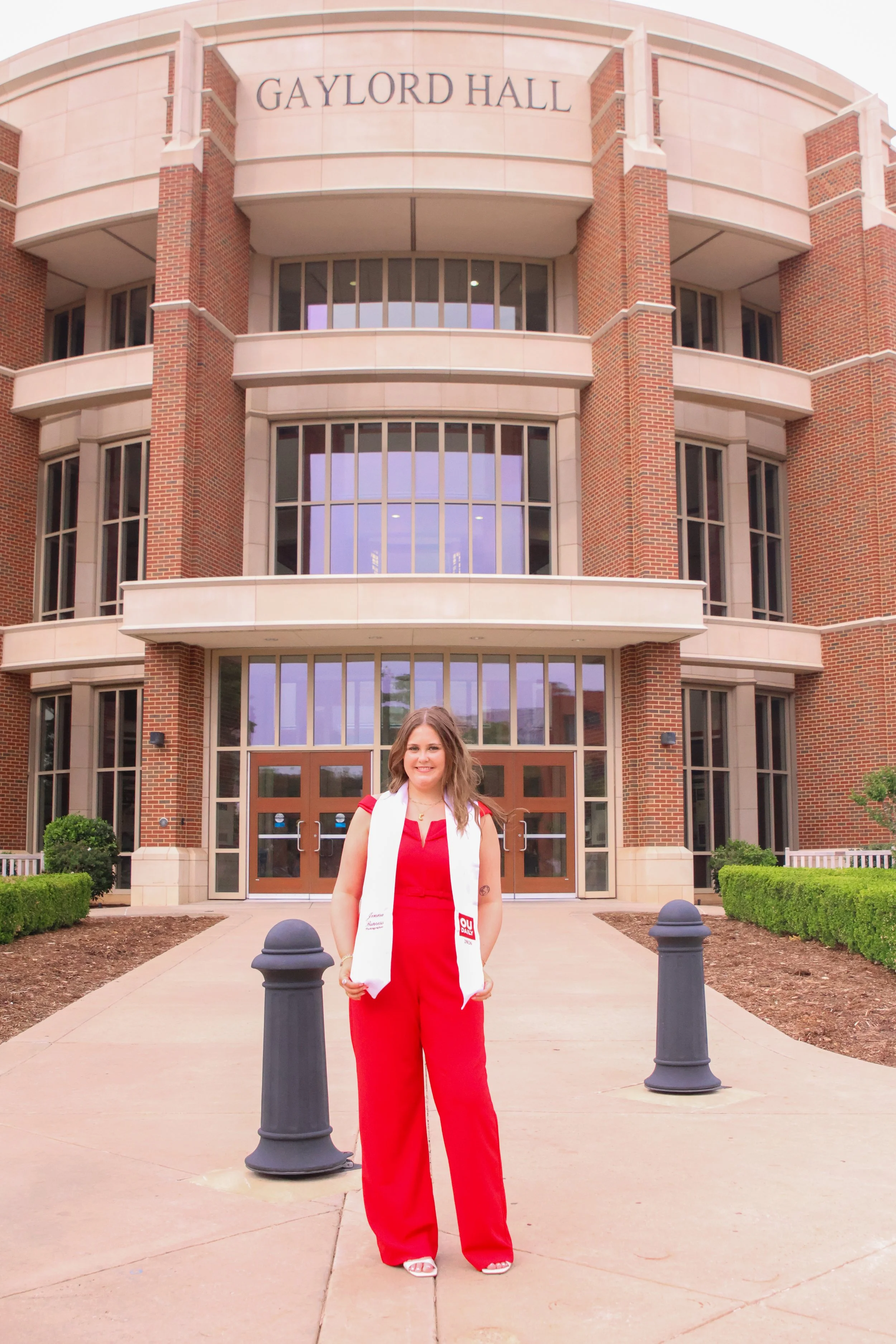 Young woman in red dress standing outside Greyard Hall building with brick walls and glass windows, surrounded by bushes and concrete pathway.