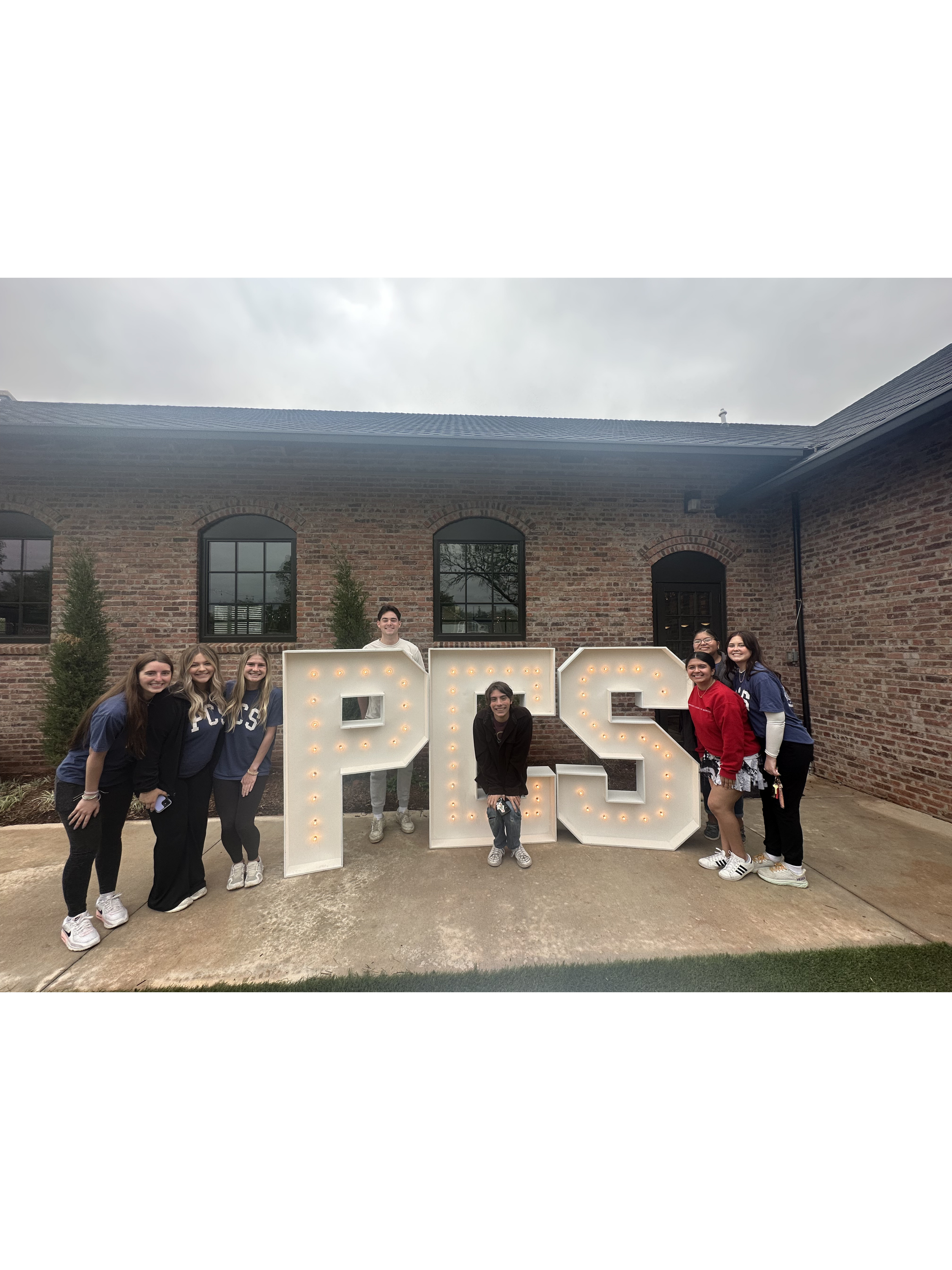 A group of eight young women and one young man standing outdoors in front of a brick building with large windows, posing behind large illuminated letters forming the number 2023, celebrating a graduation or achievement.