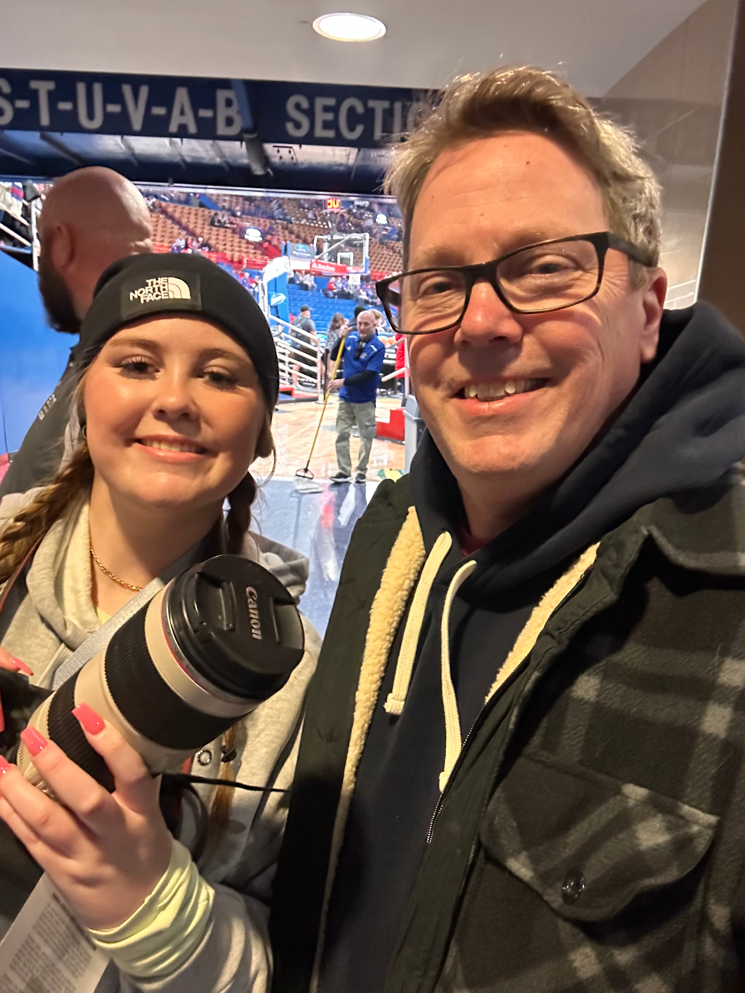 Two smiling people taking a selfie at an indoor basketball arena, with a woman holding a camera lens and a man wearing glasses, in front of a sectioned seating area and a basketball hoop.
