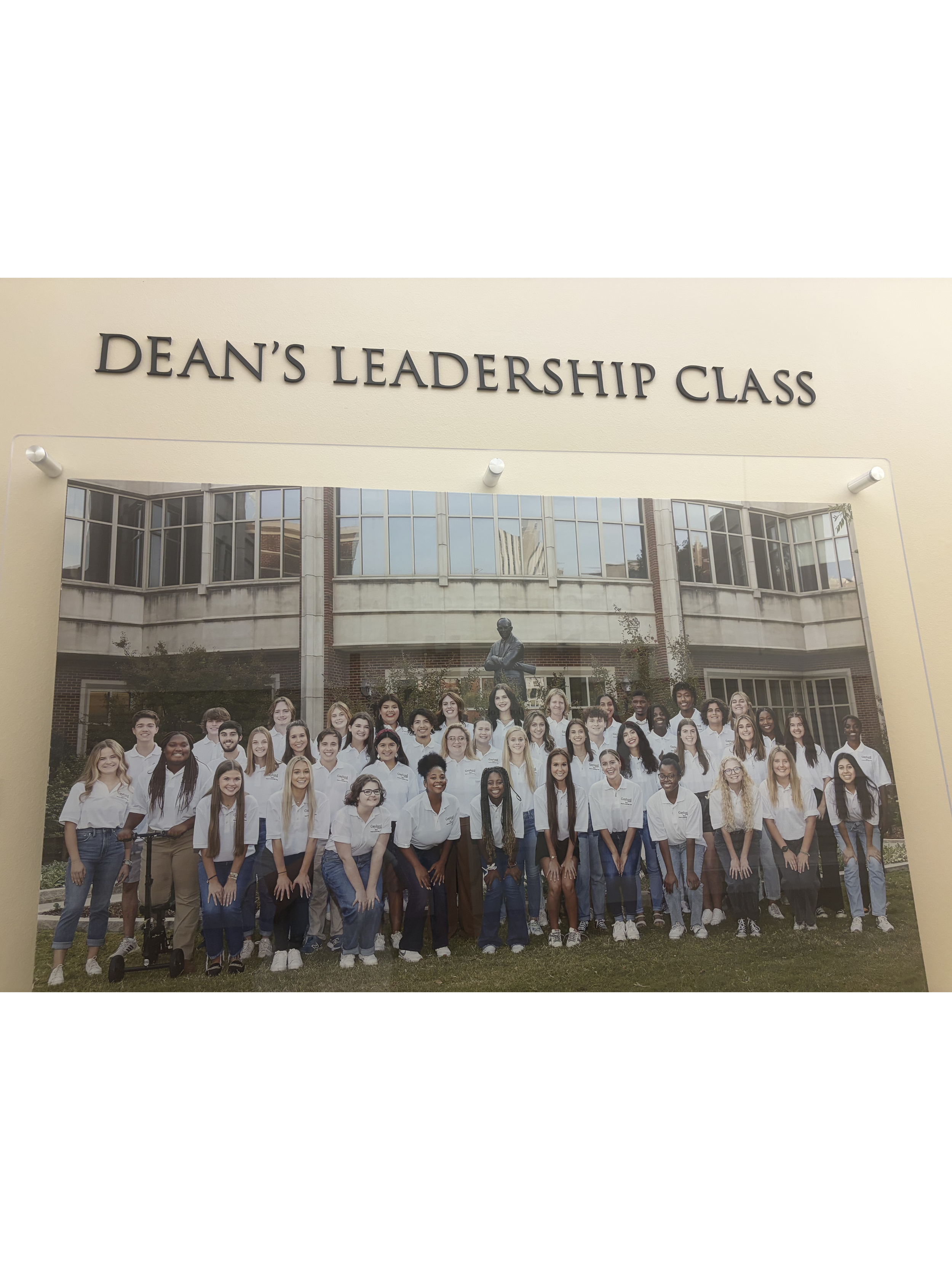 A large group of diverse students wearing white shirts and jeans, standing outside in front of a school building, posing for a group photo during Dean's Leadership Class.