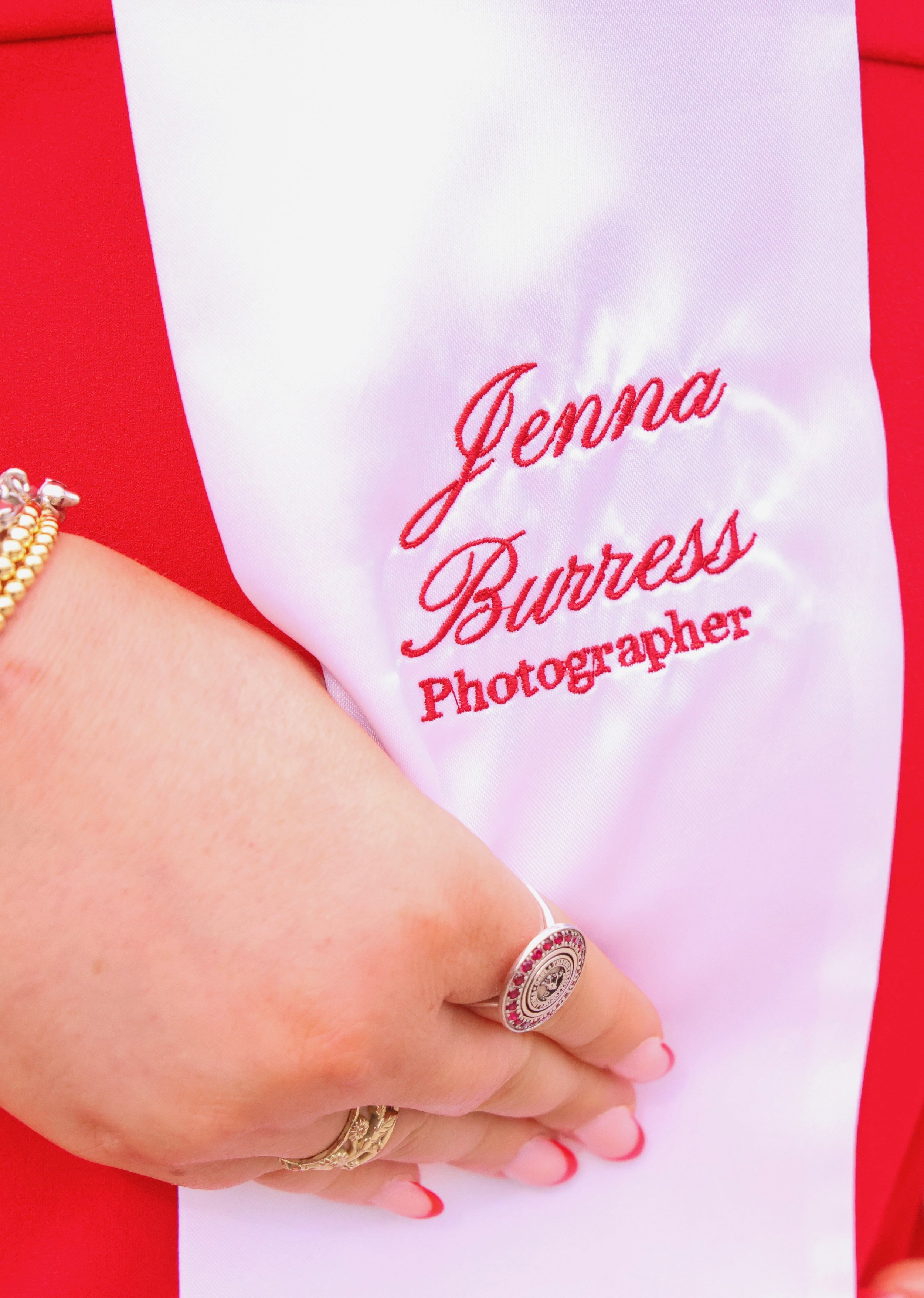 Close-up of a person's hand with rings and bracelets, holding a white cloth with red embroidered text that reads 'Jenna Burress Photographer' in front of a red background.