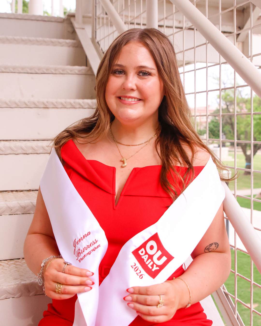 A woman wearing a red dress and a white sash that reads 'OU Daily 2026.' She has shoulder-length brown hair, is smiling, and is standing outdoors near a staircase and metal railing.