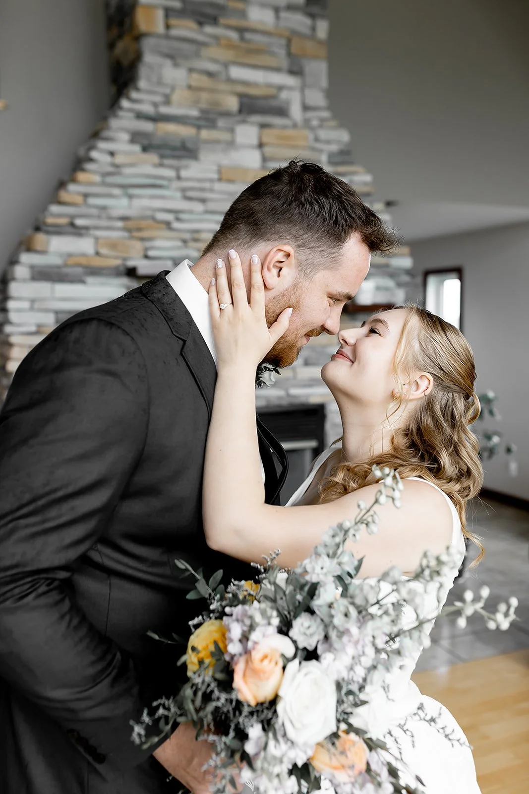 A bride and groom are close together indoors, gazing into each other's eyes. The bride has her hand on the groom's face, showing her engagement ring, and is holding a bouquet of flowers. The background features a modern stone fireplace.