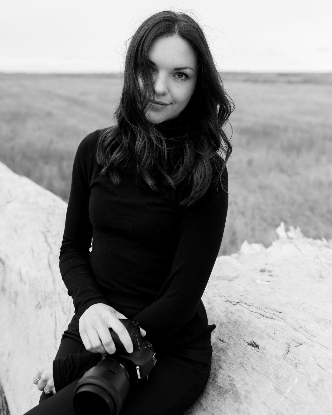 A young woman with dark wavy hair sitting outdoors on a rock holding a camera, looking at the camera with a slight smile.
