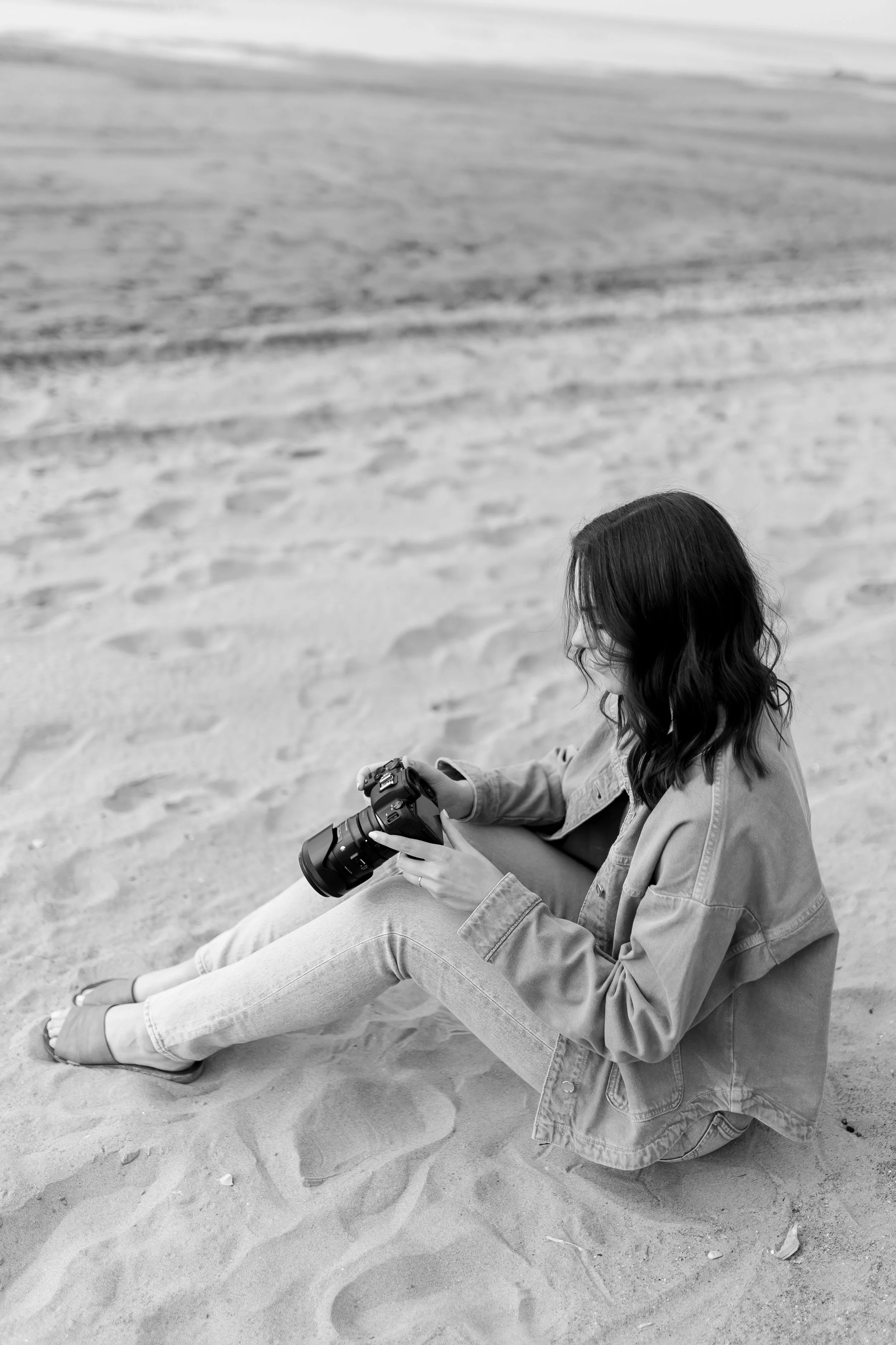 A woman with long dark hair, dressed in a denim jacket and jeans, sitting on sandy ground at the beach, holding a camera and looking at it. The background features sand with some footprints.